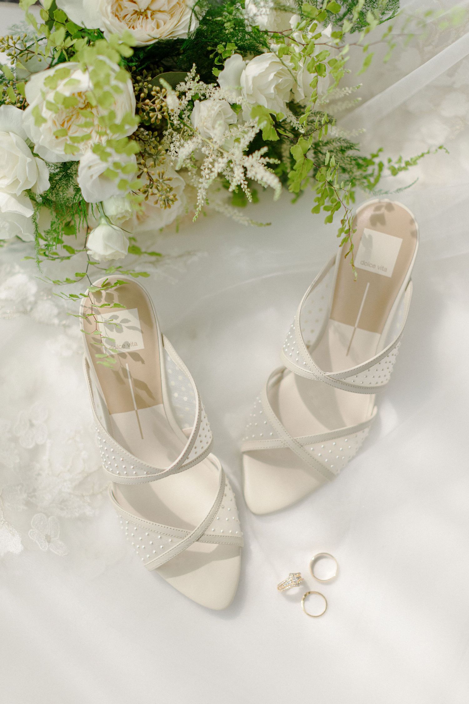 White bridal heels and wedding rings styled with fresh flowers on a white surface.