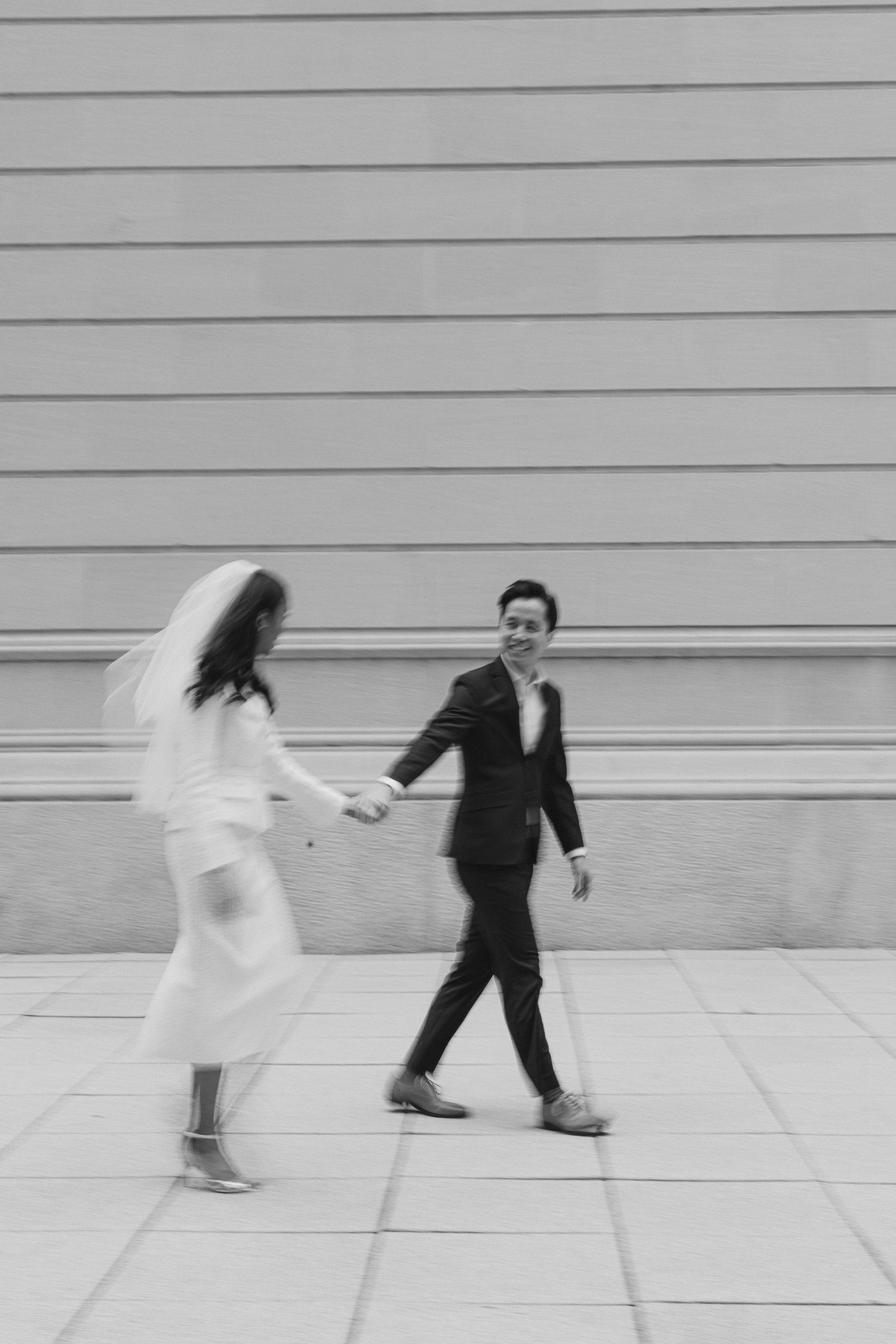 Artistic black and white photo of newlyweds dancing playfully against minimal wall background.