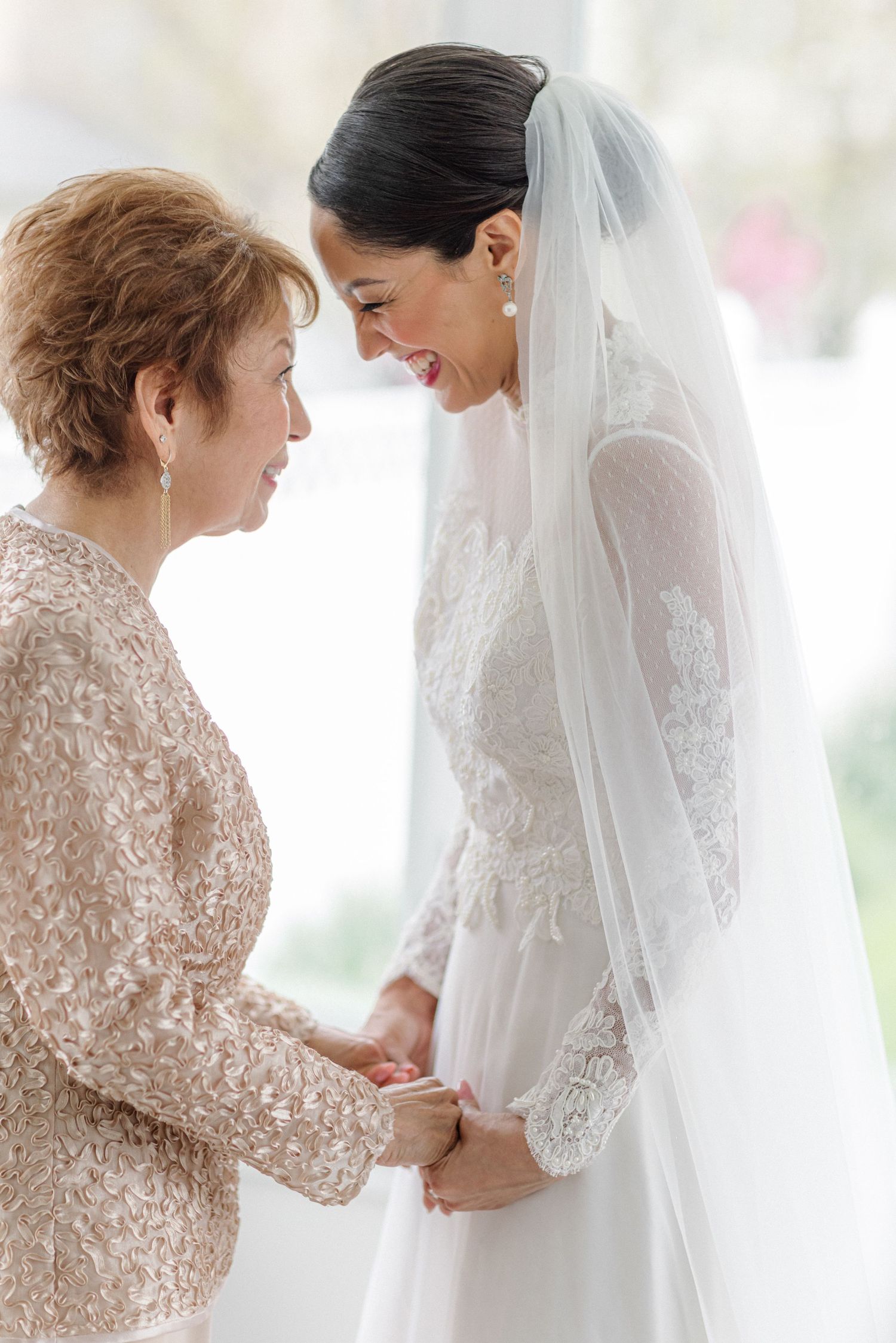 A tender moment between a bride in lace gown and woman in champagne dress during wedding preparations.