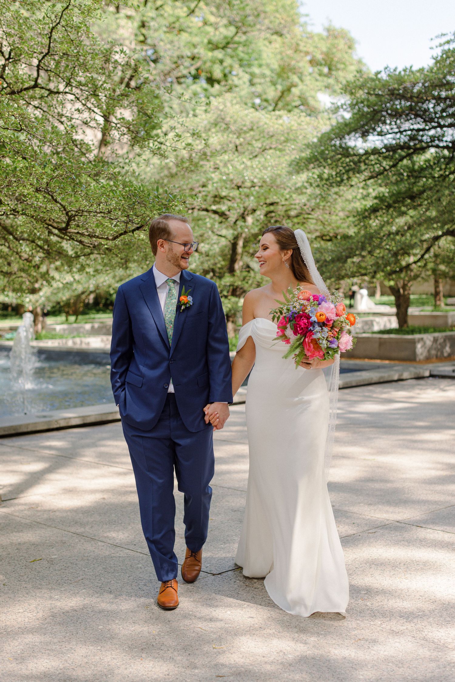 A happy couple walking through a park with fountain, carrying bright floral bouquet in wedding attire.