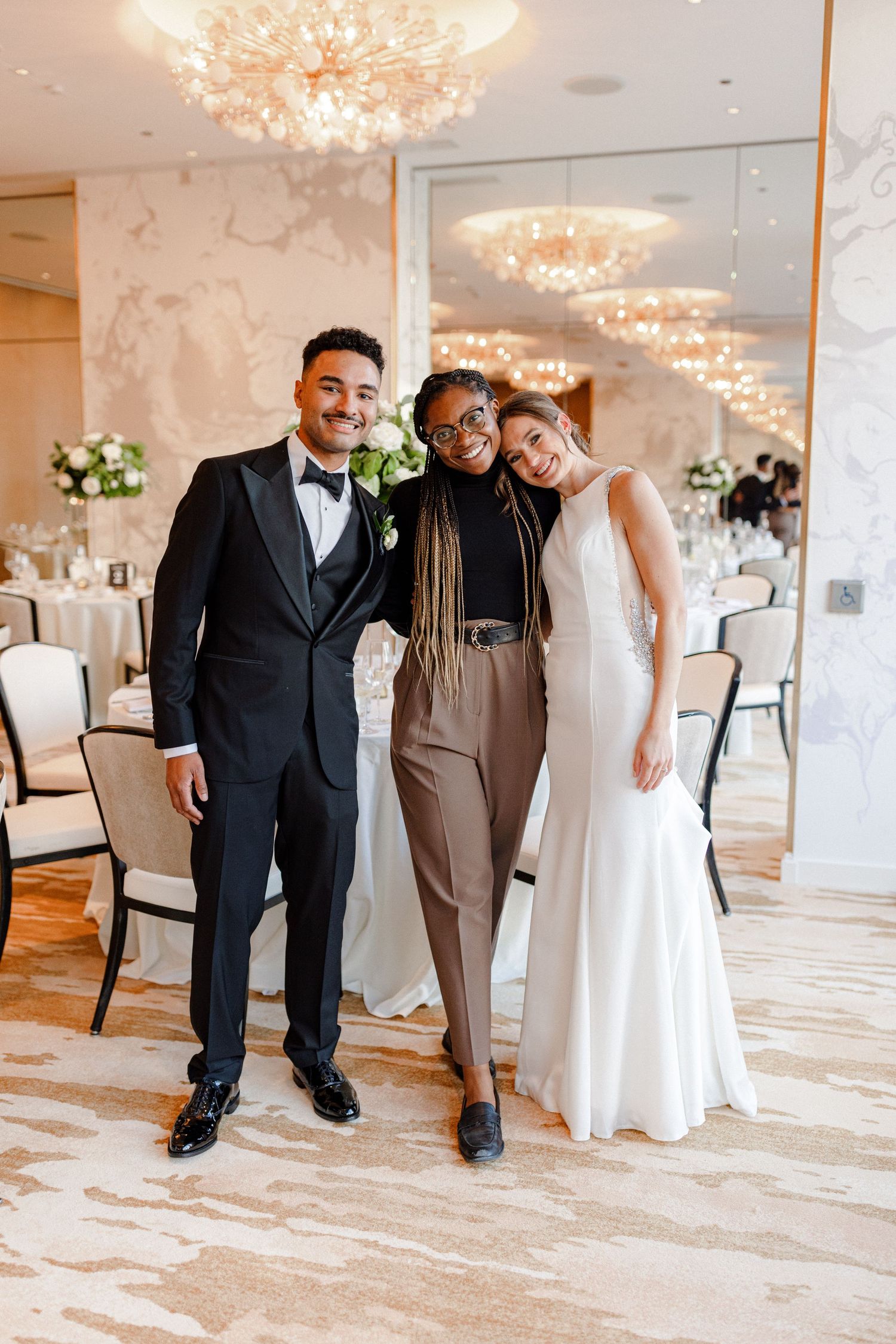 Three people in formal attire pose in elegant ballroom with chandeliers.