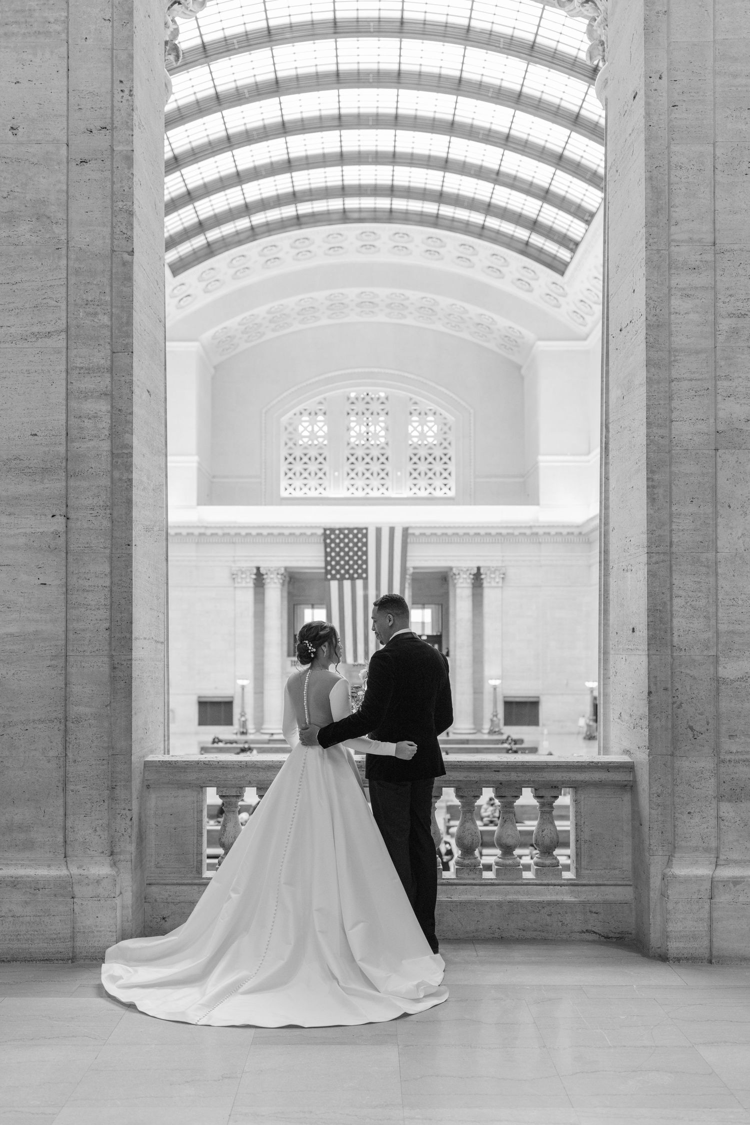 Classic black and white wedding photo of couple overlooking grand hall with American flag.
