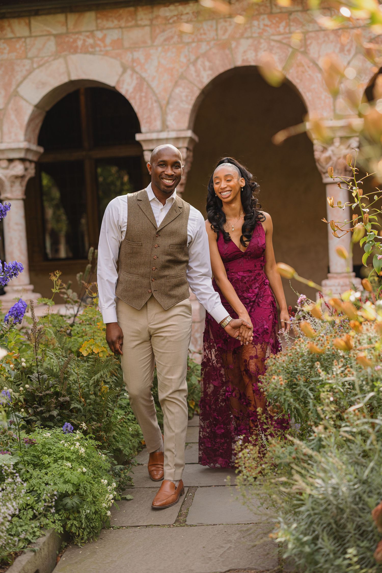 A couple walking together in an outdoor garden setting with stone arches and greenery in the background.