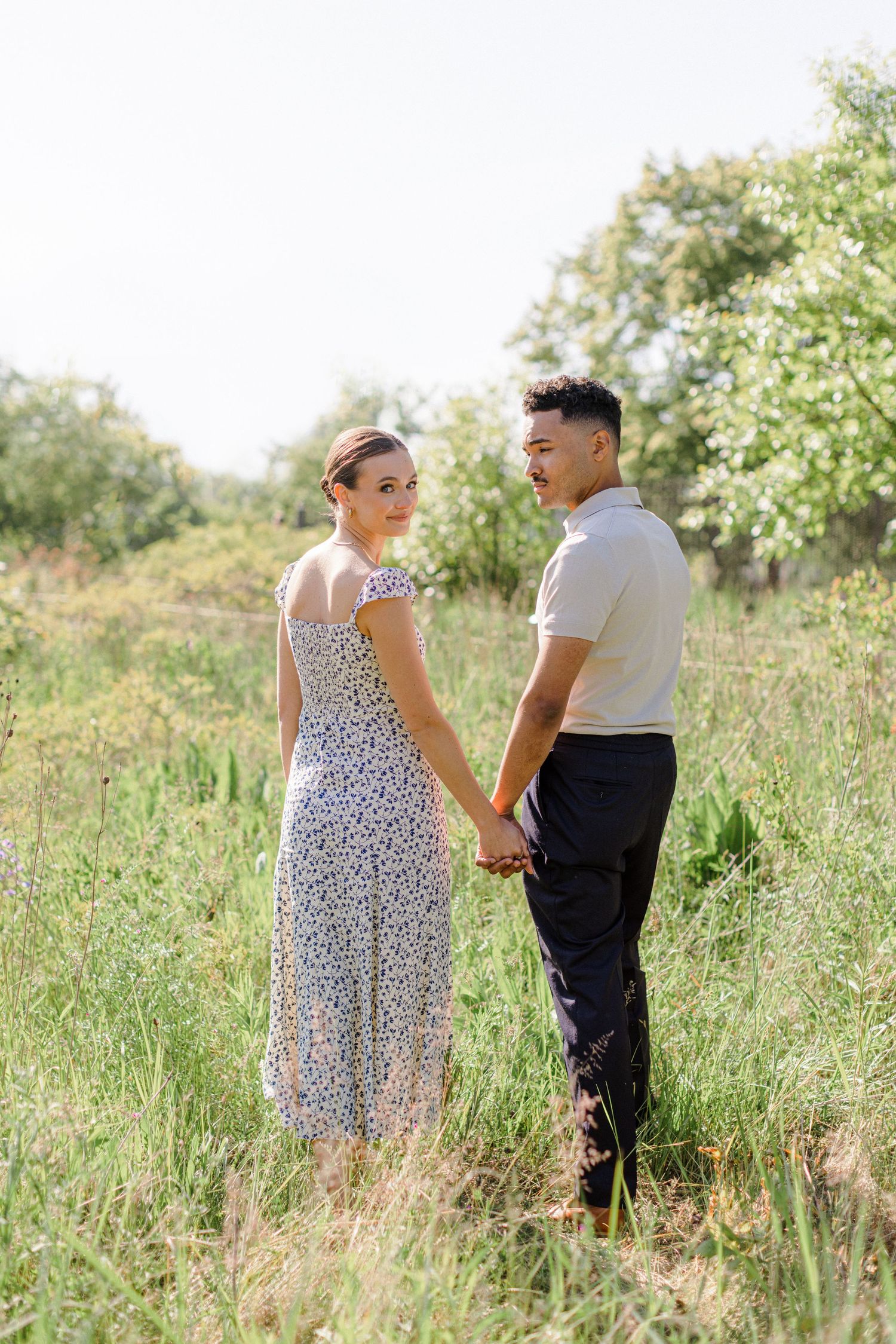A couple holds hands in a sunlit meadow with tall grass and trees in the background.
