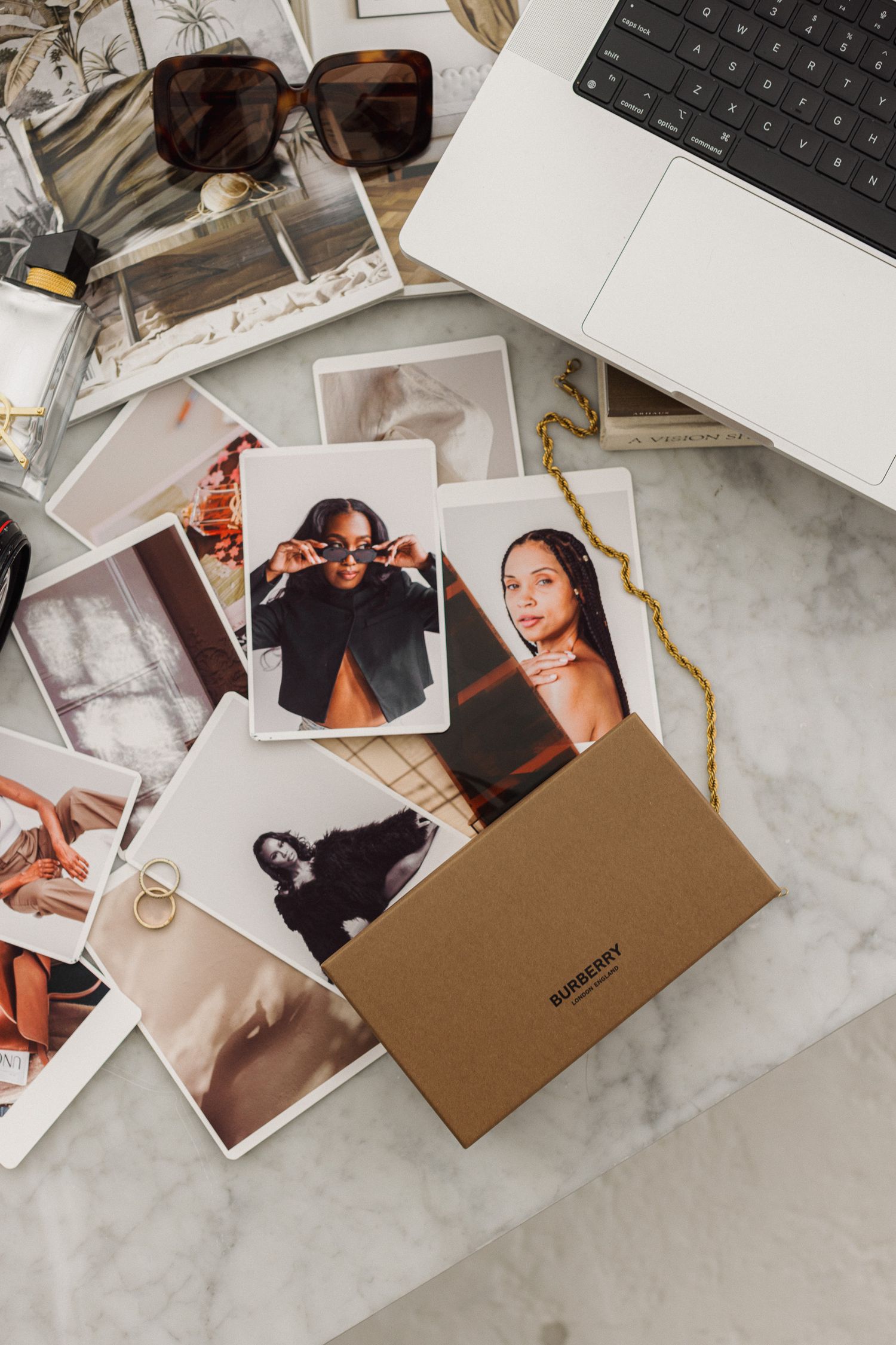 Scattered polaroid photos and sunglasses lay beside a laptop on a marble surface with a brown envelope.