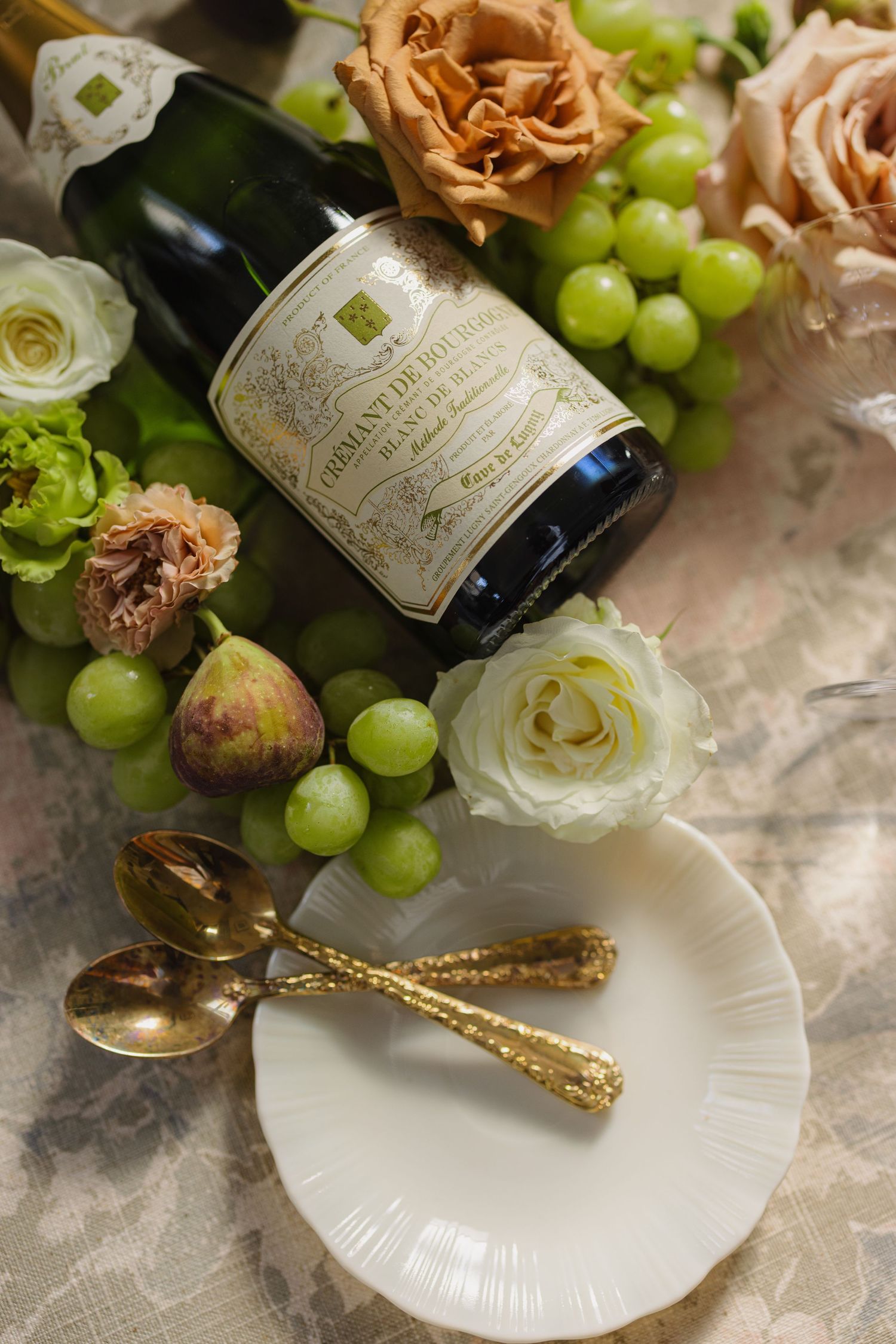 Elegant table setting with champagne bottle, white roses, green grapes, and gold utensils on white plates.