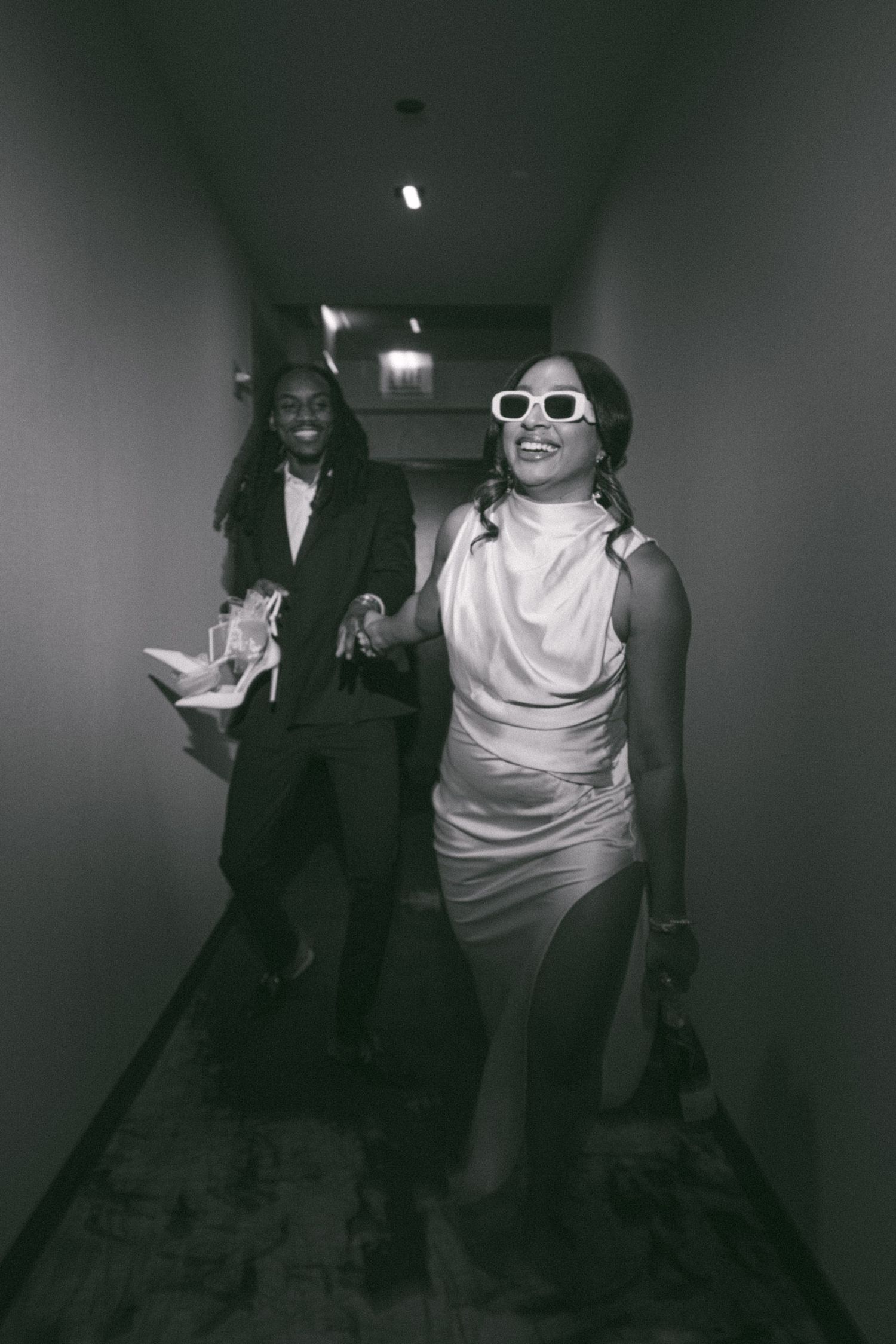 Two people laughing and walking down a dimly lit hotel hallway in formal wear.