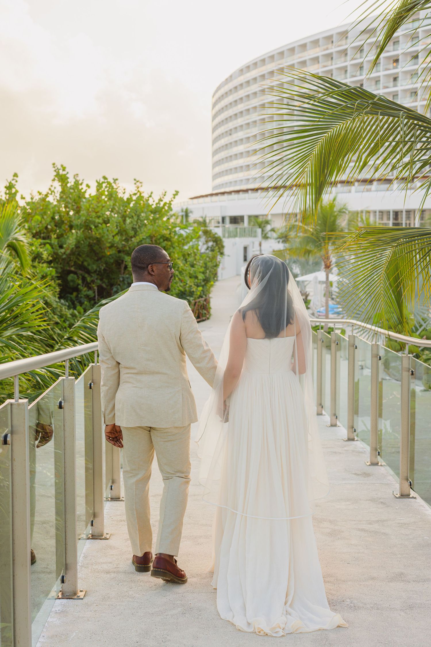 Newlywed couple walking on resort pathway with palm trees and modern hotel building in background at sunset.