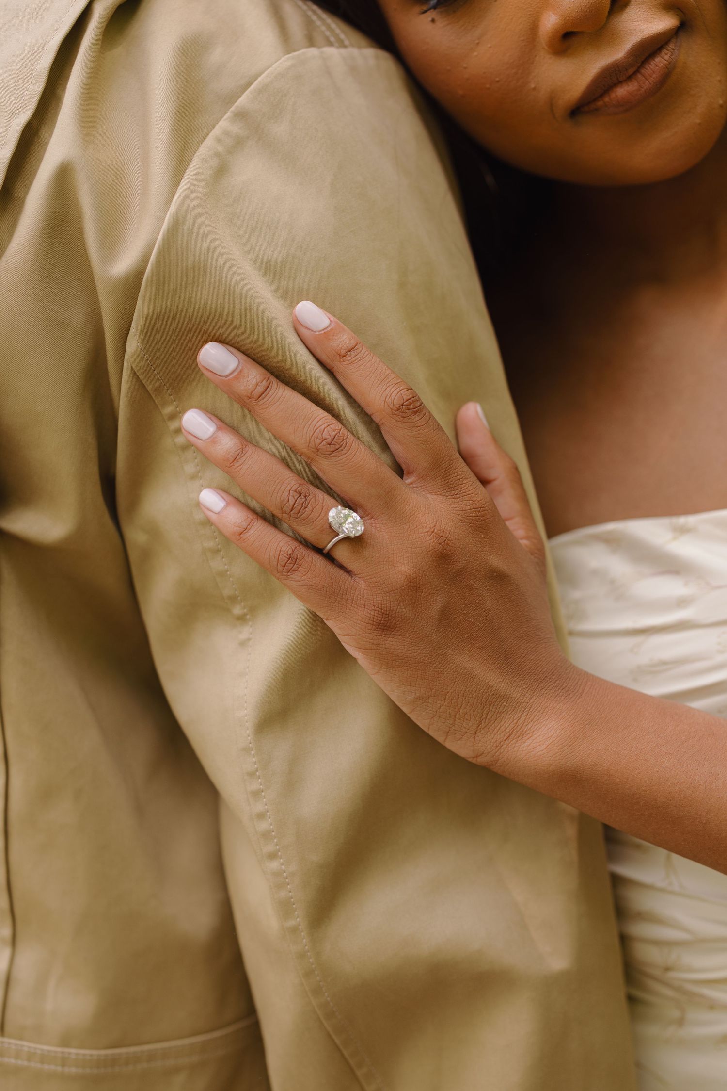 A hand wearing a white manicure and diamond engagement ring is shown against a beige fabric background.