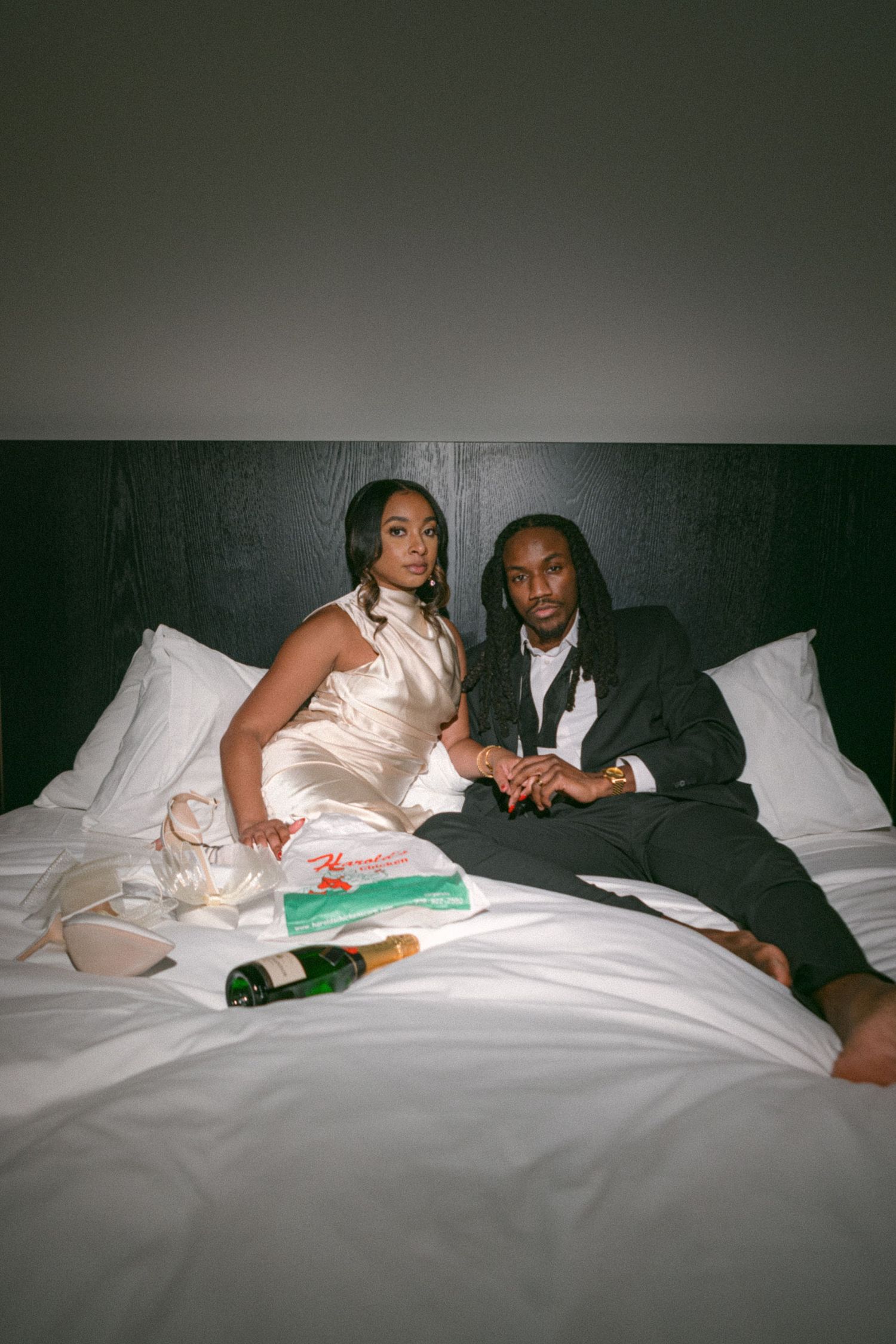 A newlywed couple relaxing on white hotel bedding with champagne.