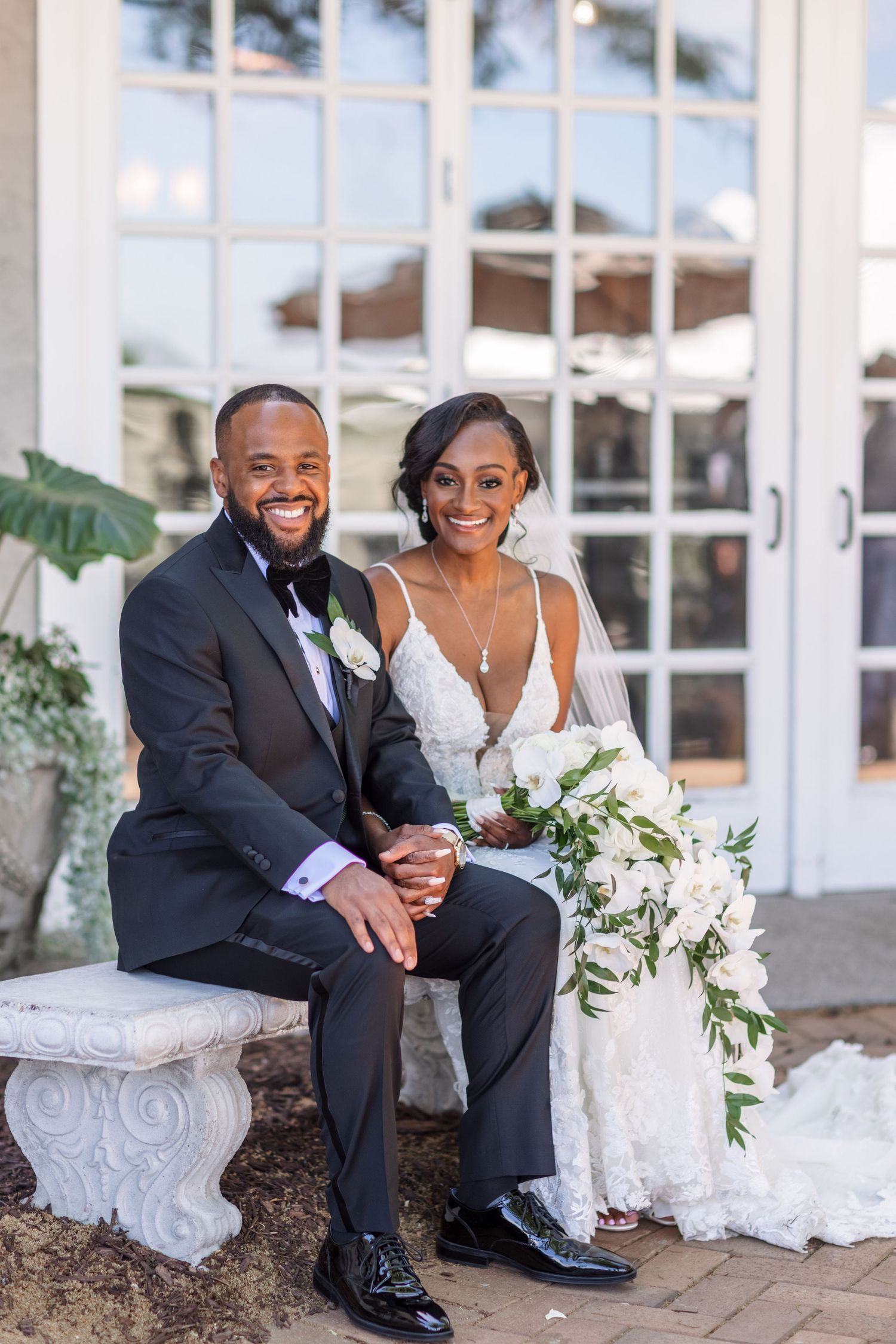 A couple sits together on a stone bench in formal wedding attire with a floral bouquet.