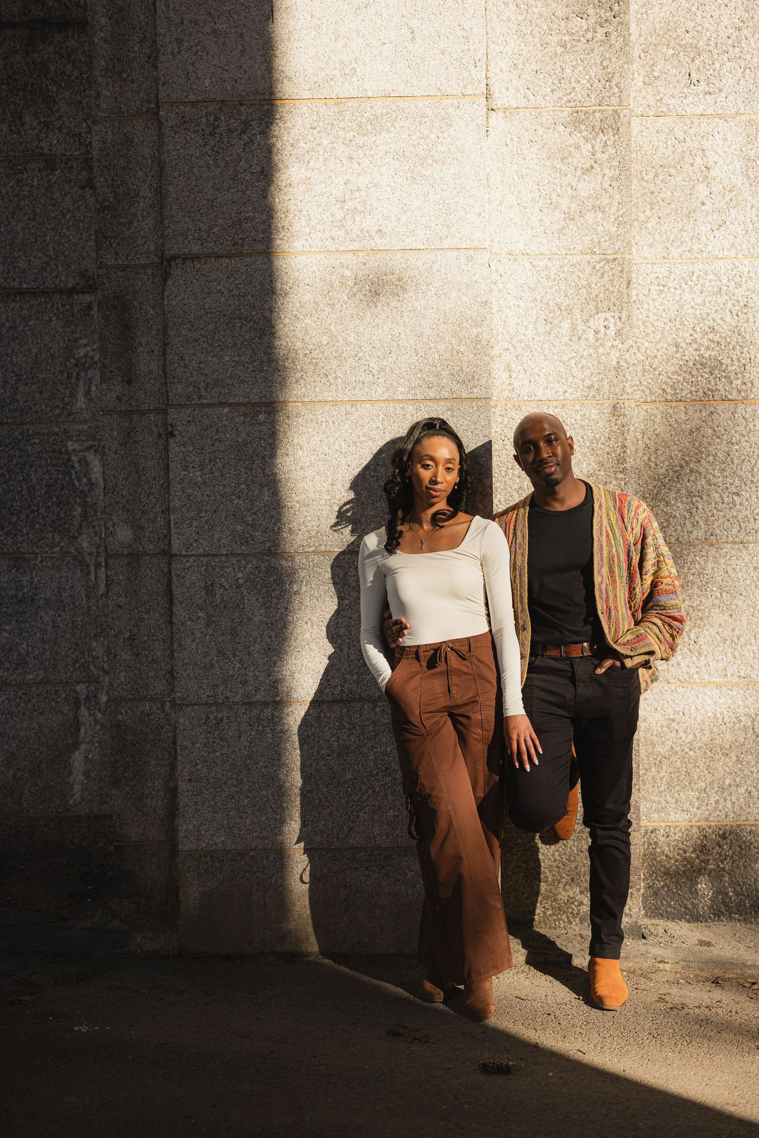 Two people in stylish casual wear pose against a sunlit concrete wall creating dramatic shadows and contrast.