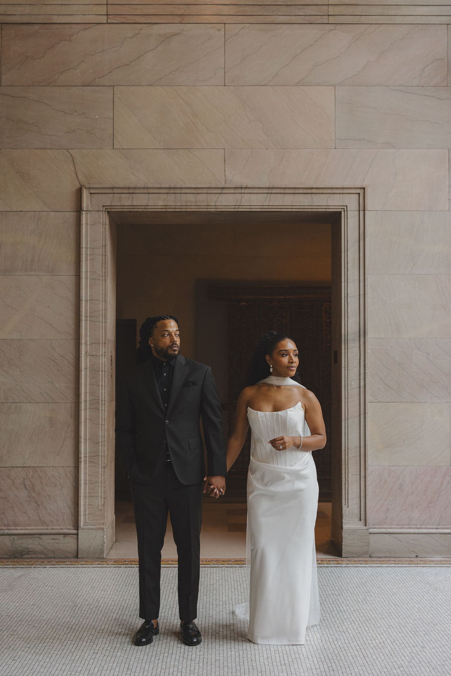 A couple in wedding attire stands together in a minimalist marble hallway for artistic portraits.