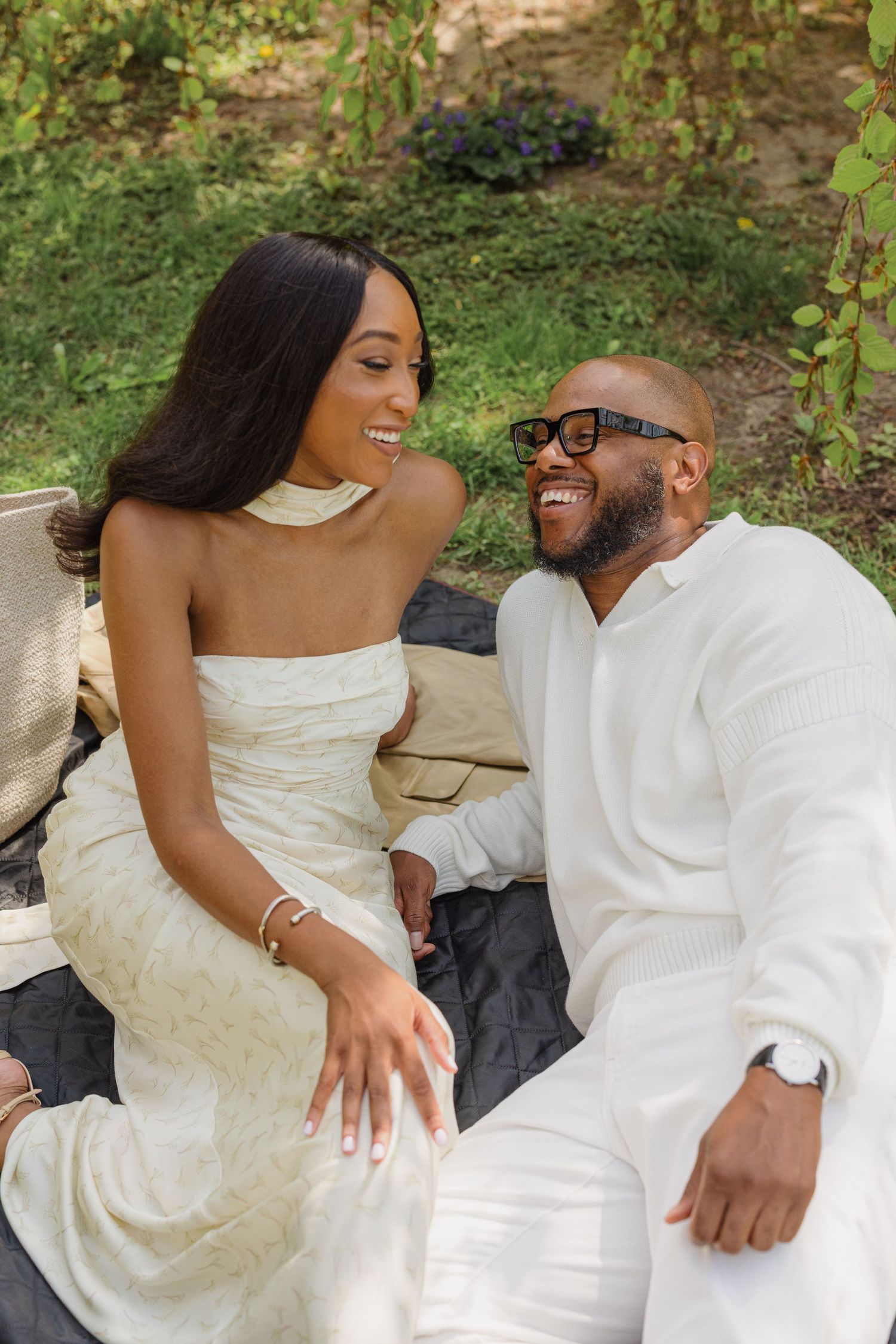 A couple in white attire share intimate moments while sitting together outdoors in a natural setting.