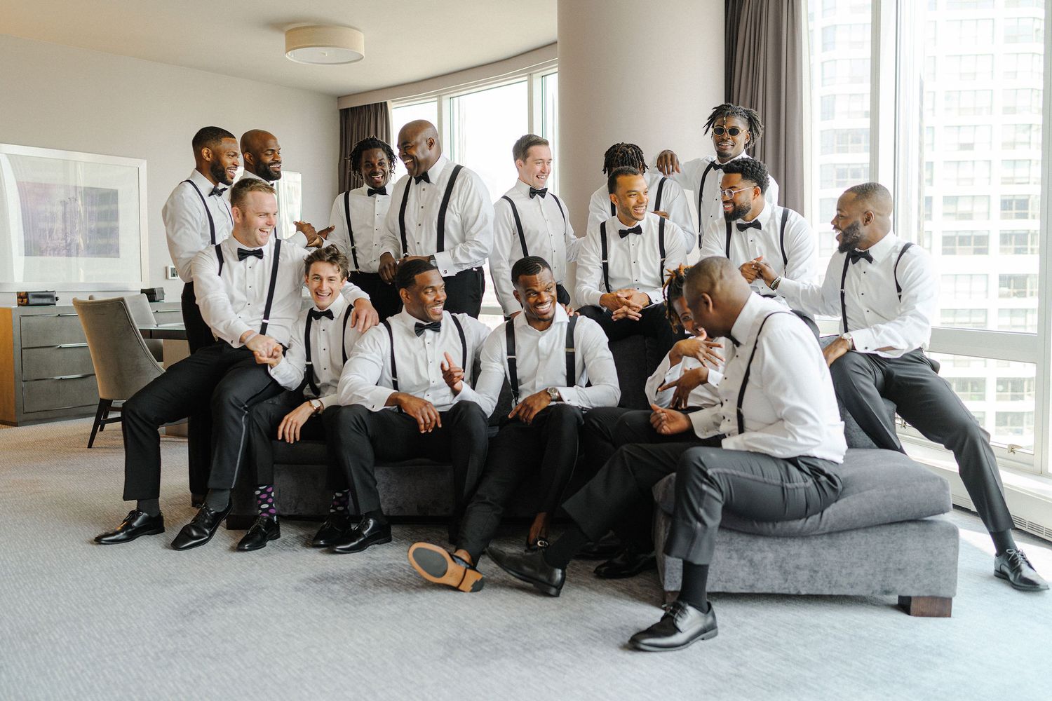 Groomsmen in matching black and white attire with suspenders posing together on a gray couch in a modern room.