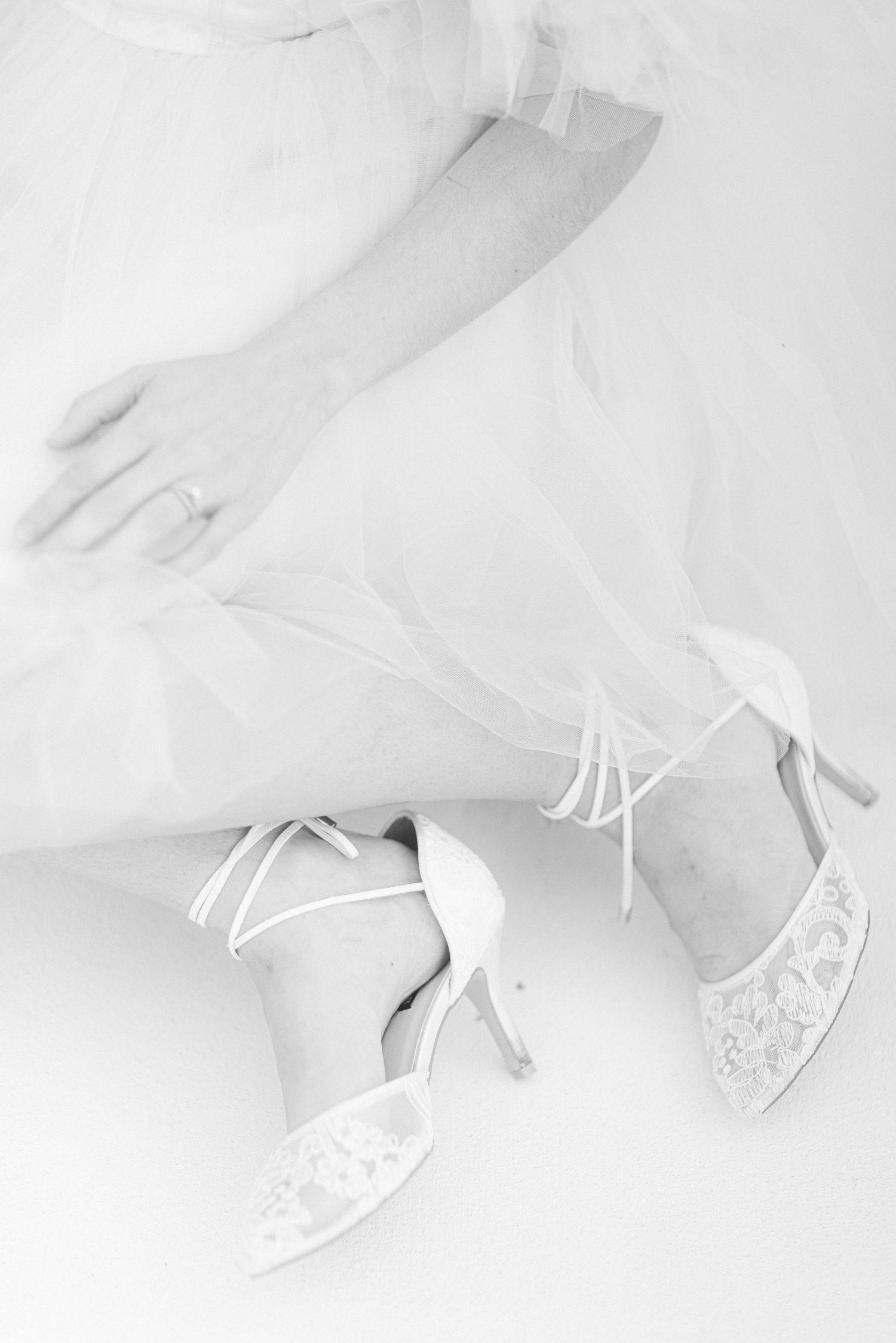White lace wedding shoes with ankle straps seen against flowing dress fabric in soft lighting.