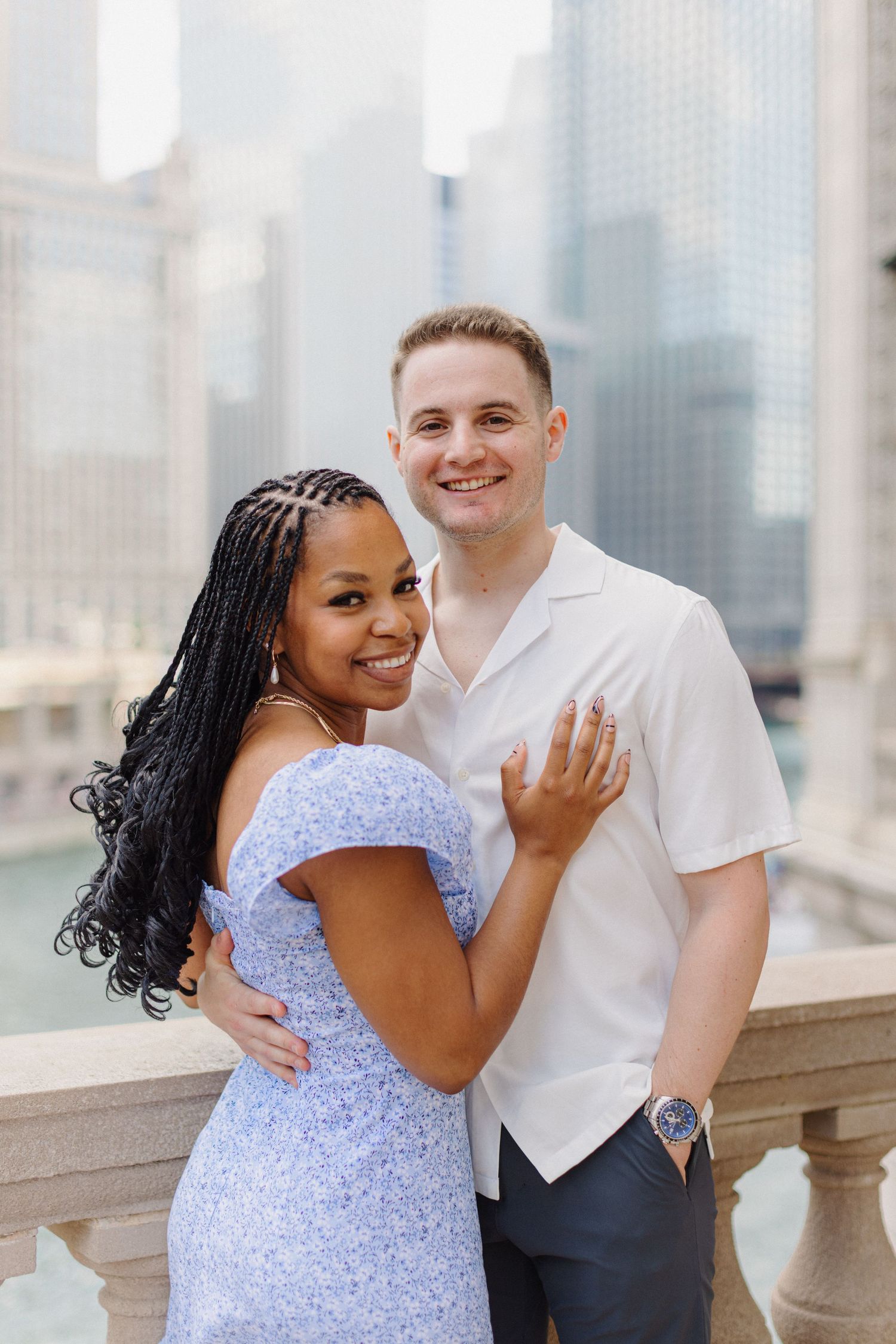 A couple poses together on a city balcony, showing off an engagement ring.