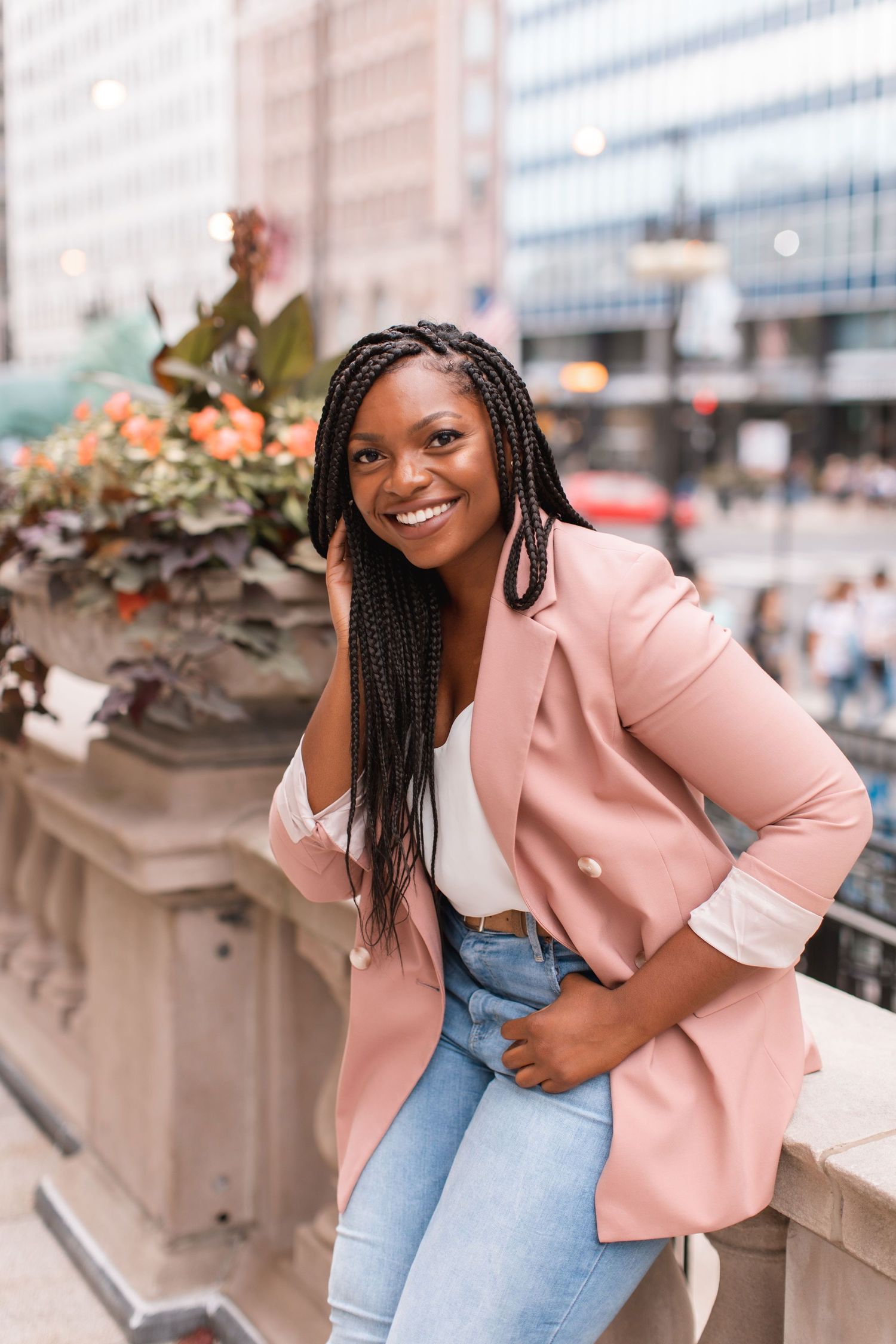 Woman in pink blazer and jeans posing outdoors by urban planter.