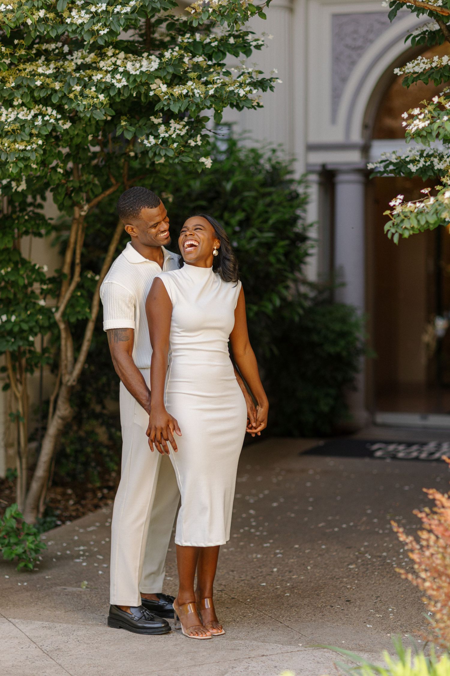 Couple in elegant white outfits share a laugh while standing in a garden archway.