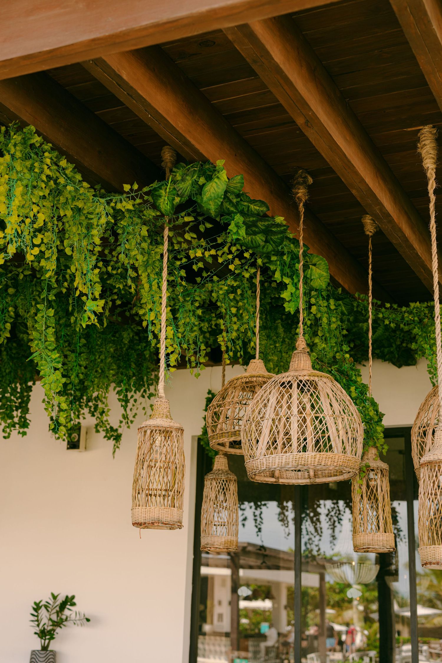 Rustic woven rattan pendant lights hang beneath wooden beams adorned with cascading green vines in an outdoor patio.