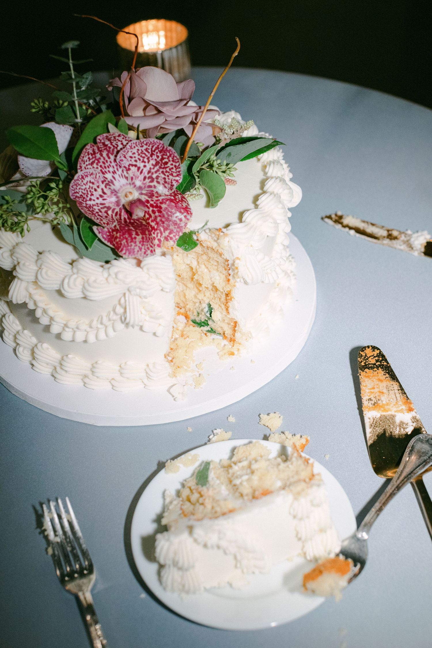 A white wedding cake decorated with pink orchids and greenery sits sliced on a table.