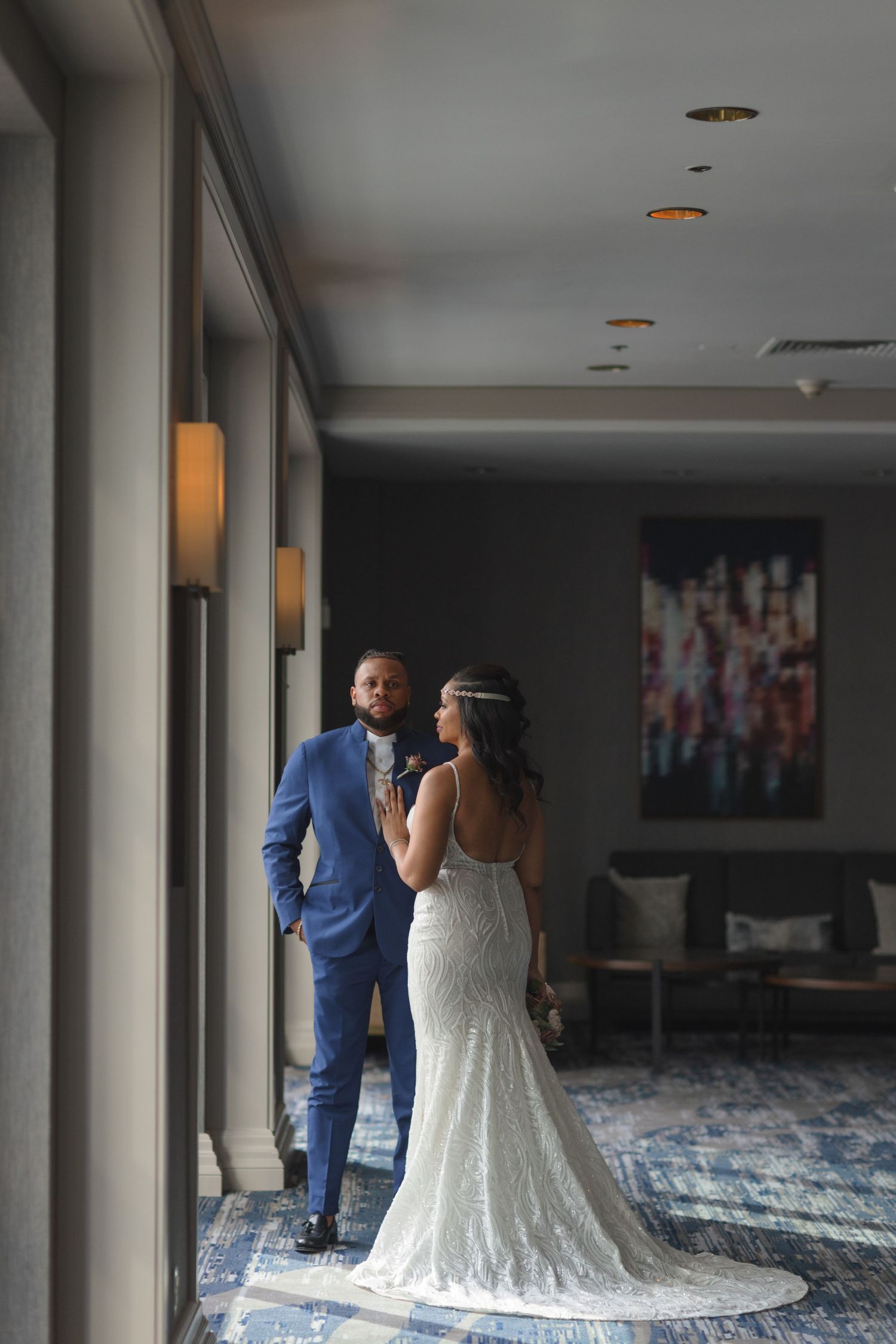 A couple in elegant wedding attire sharing a private moment near large windows in a modern venue.