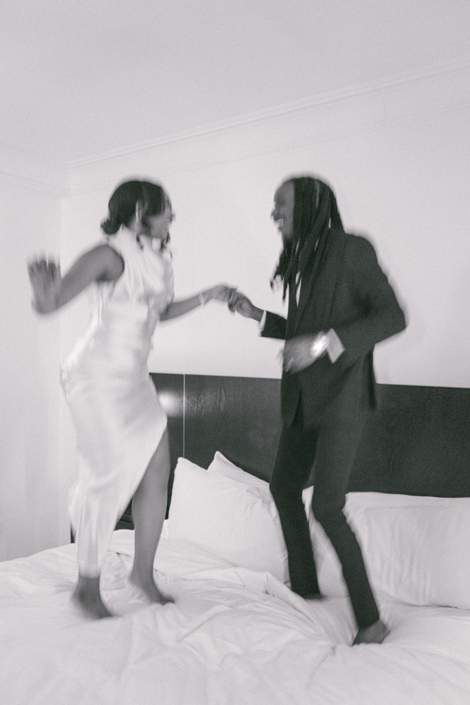 Playful black and white photo of couple dancing on hotel bed.