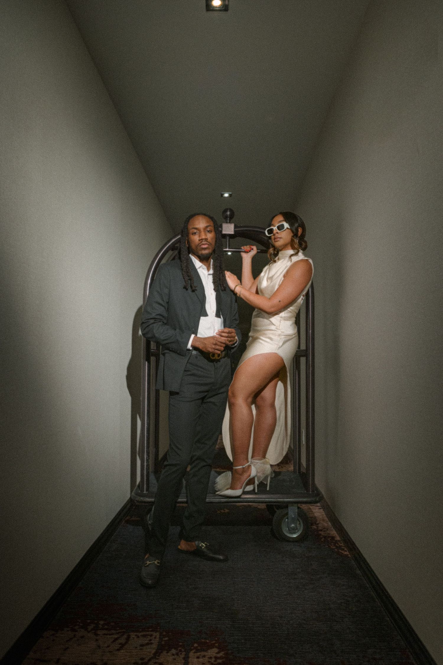 Stylish couple posing with hotel luggage cart in dimly lit hallway.