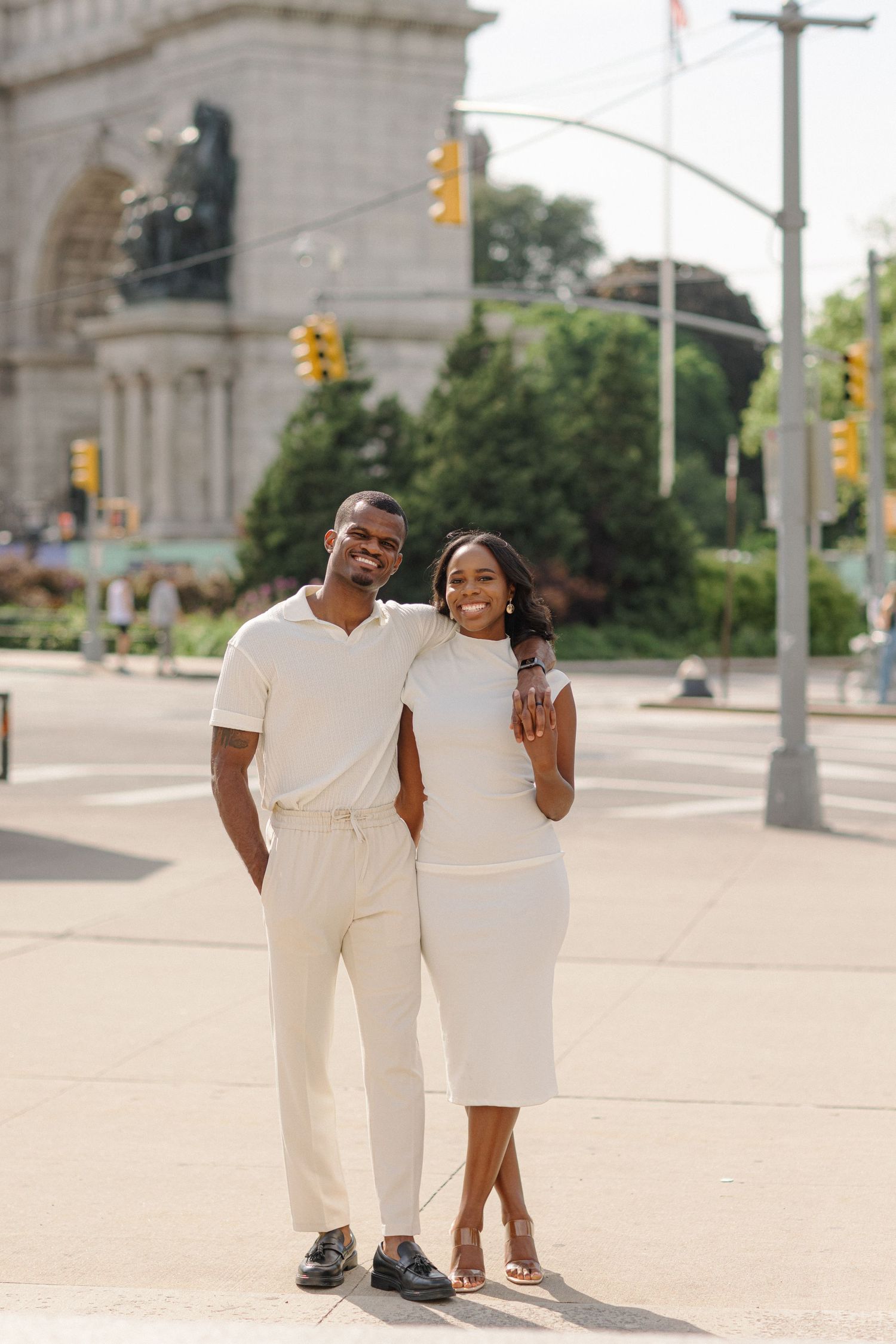 A stylish couple in coordinating white outfits pose together on a city street with traffic lights and architecture in background.