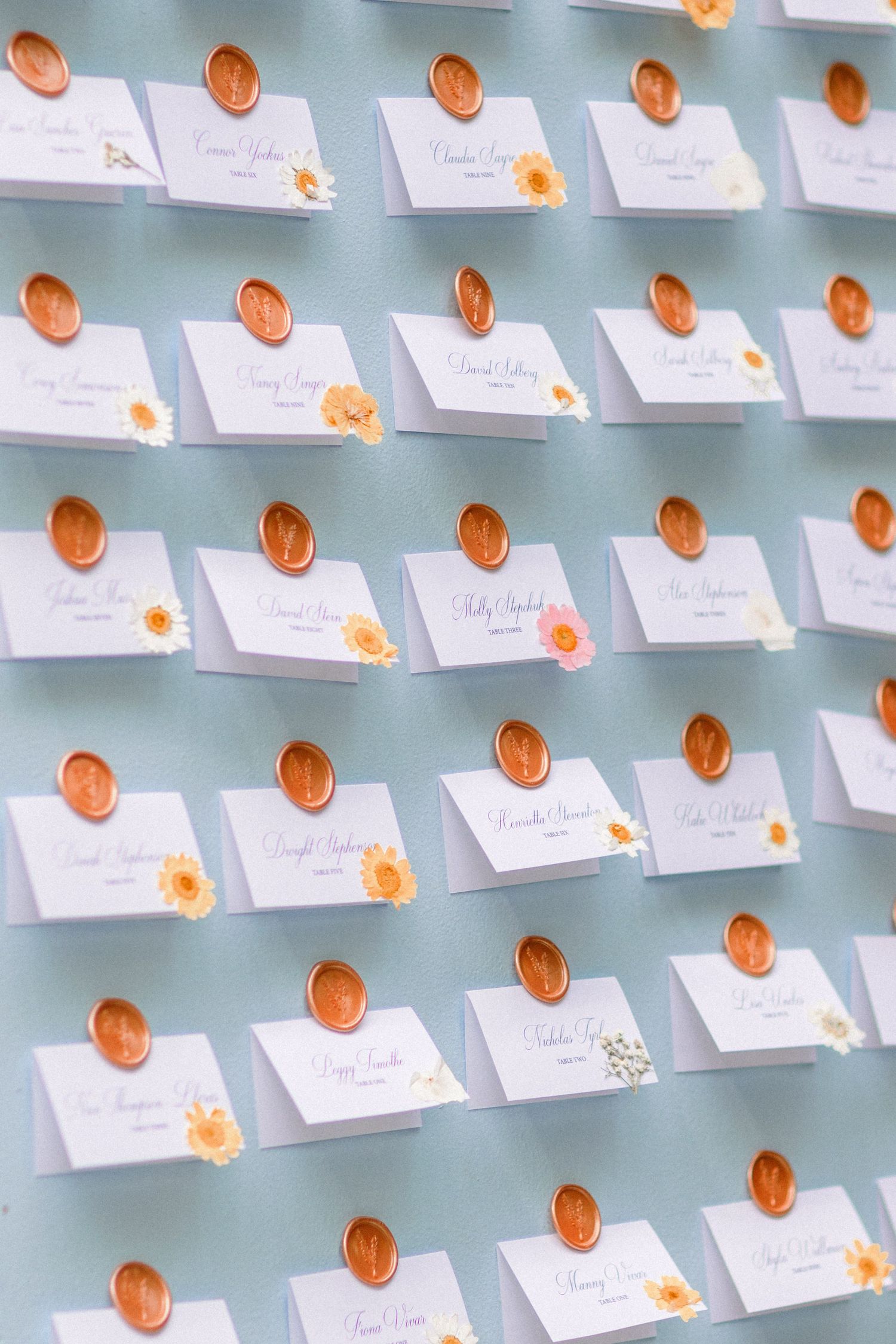 Wedding escort cards with orange wax seals arranged in neat rows on a blue-grey background.
