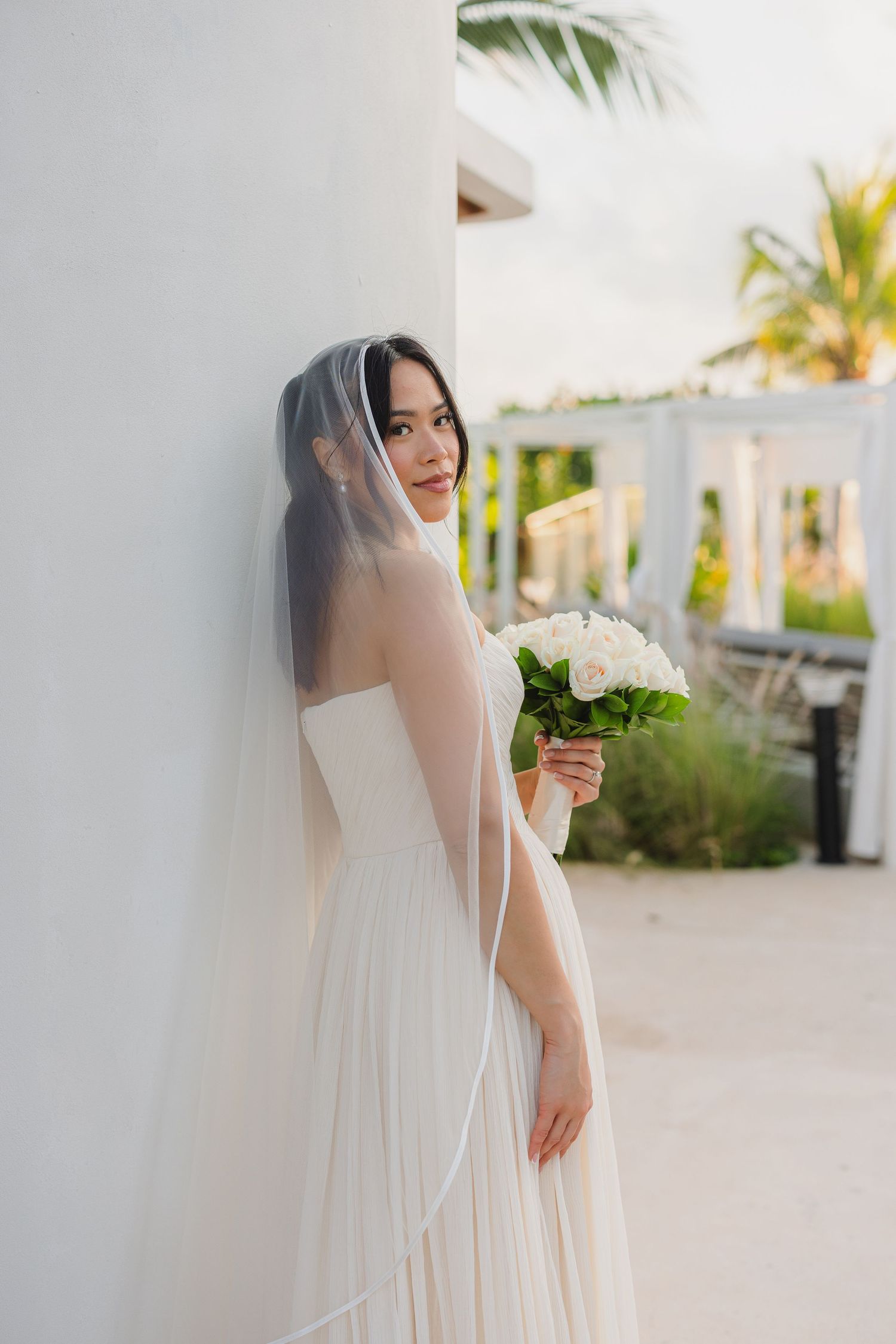 A bride in a flowing white wedding dress and veil holds a bouquet of white flowers against a tropical outdoor setting.