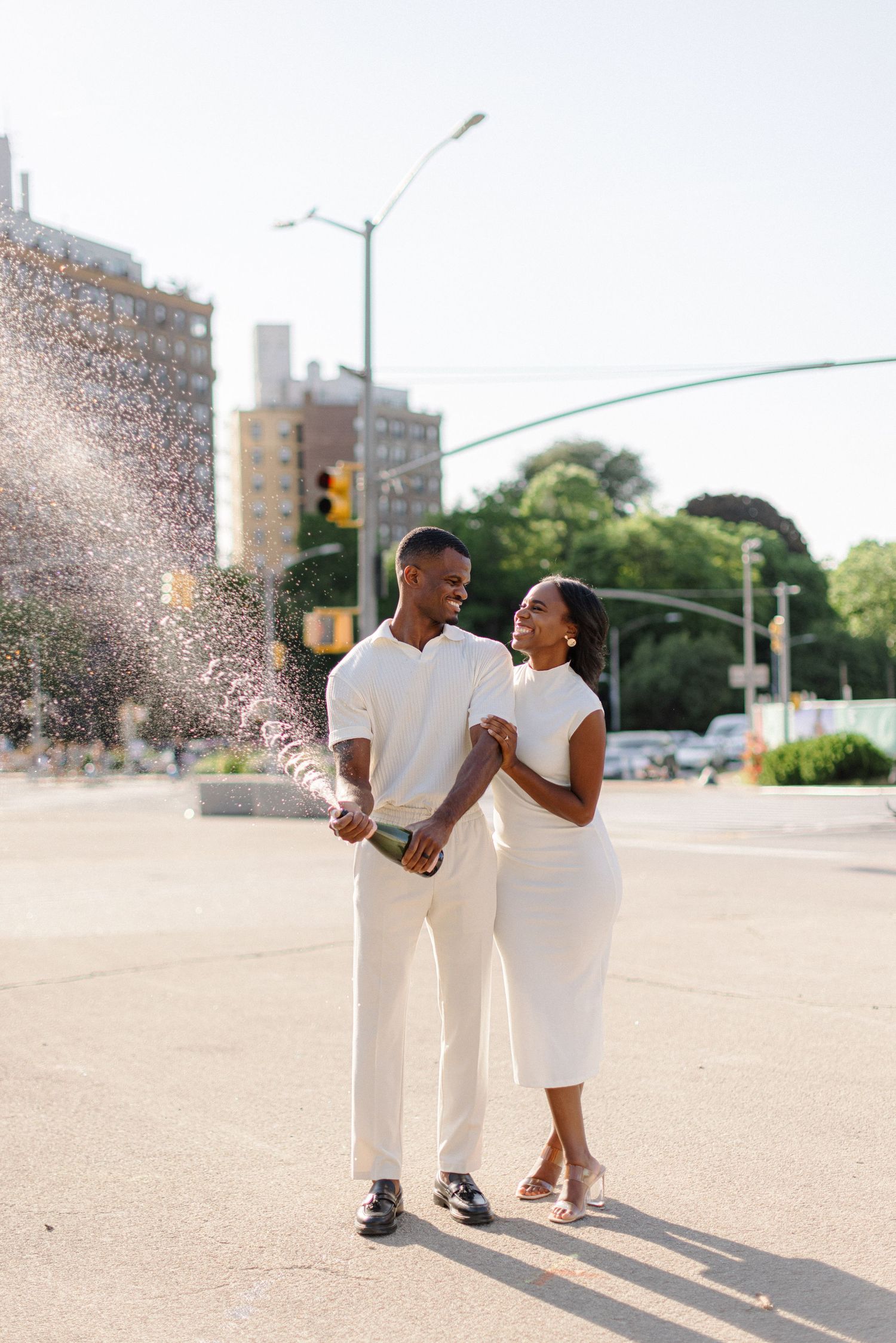 A couple in white outfits laughs together on a sunny city street.
