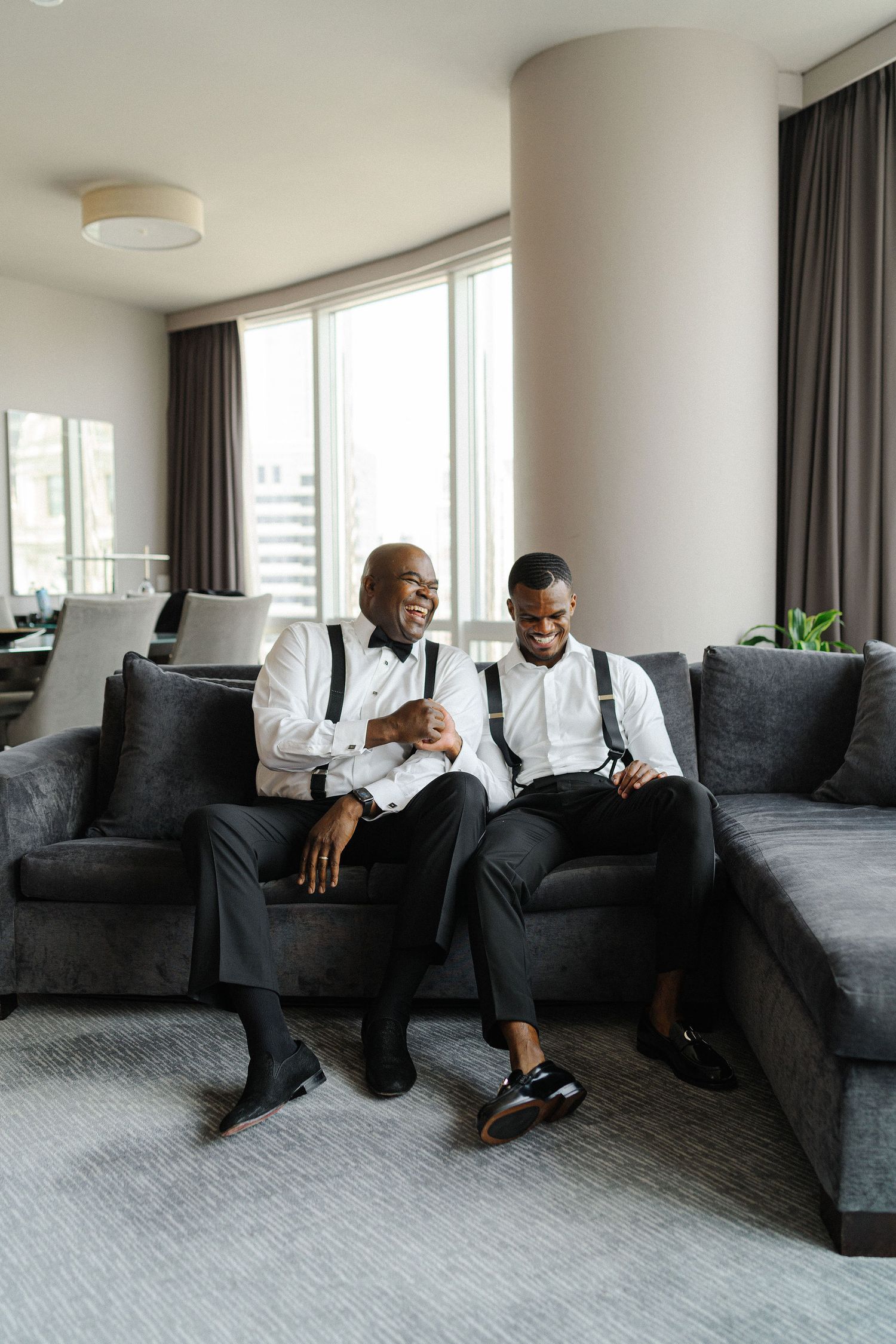Two people in formal attire with suspenders and bow ties sharing a laugh on gray sectional sofa.