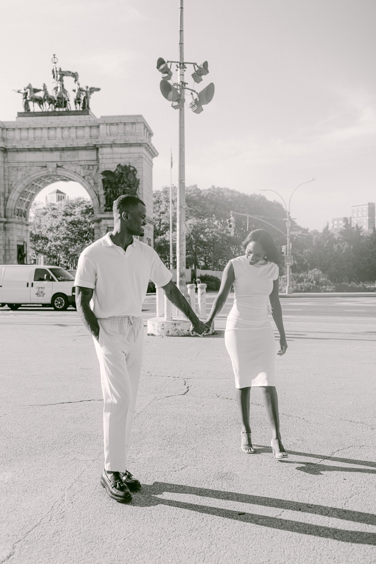 A couple in white outfits holds hands while walking near a grand archway.