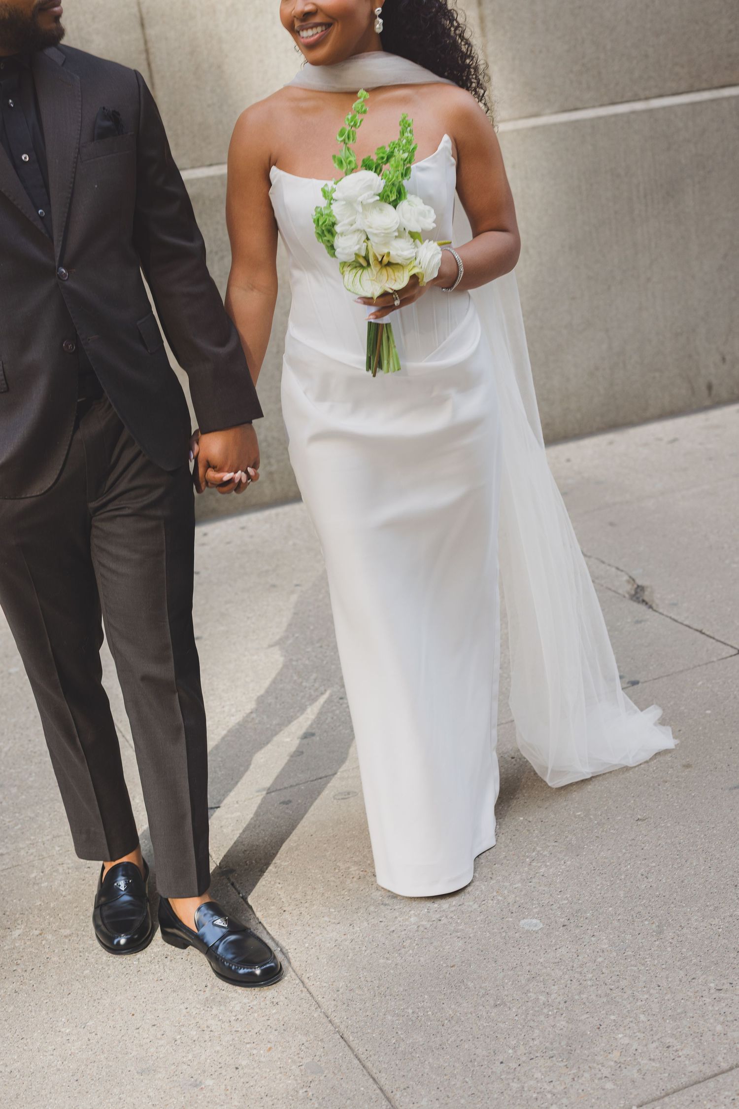 A bride in a white wedding dress holds a green and white bouquet while standing next to someone in a dark suit.