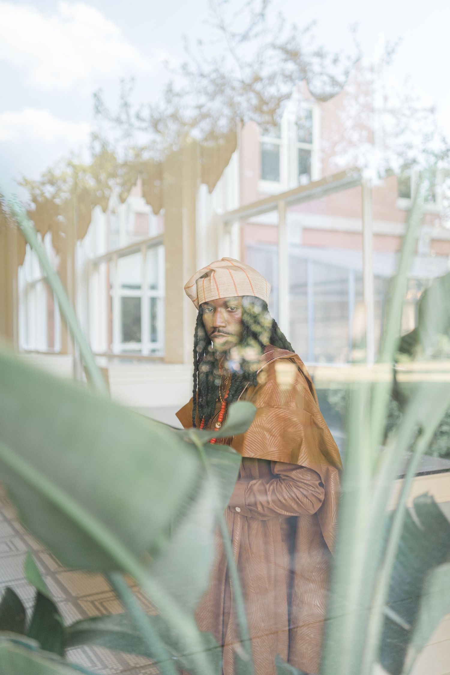 A person in earth-toned clothing poses thoughtfully among green plants.