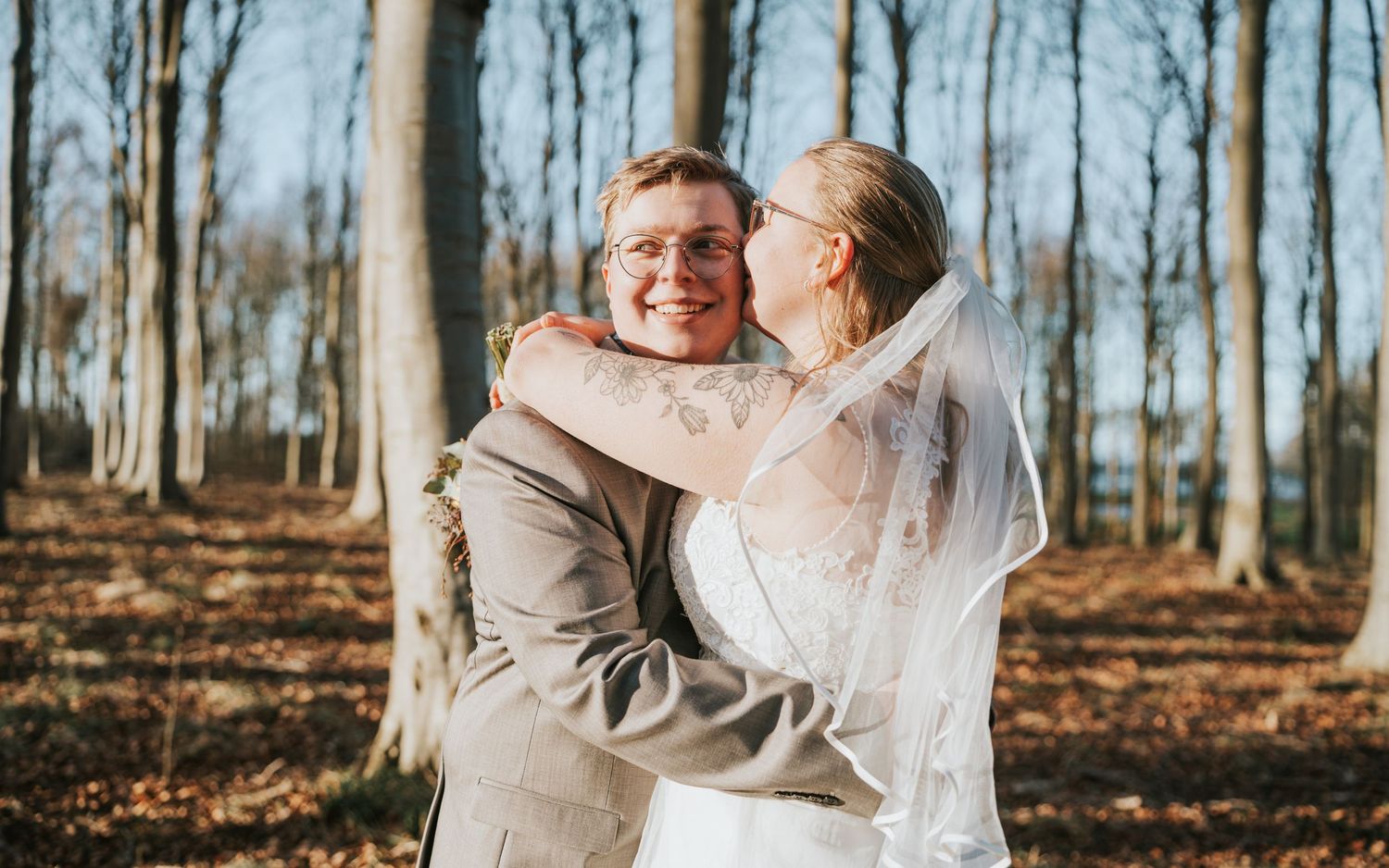 A happy couple embraces in a wooded autumn setting wearing elegant wedding attire.