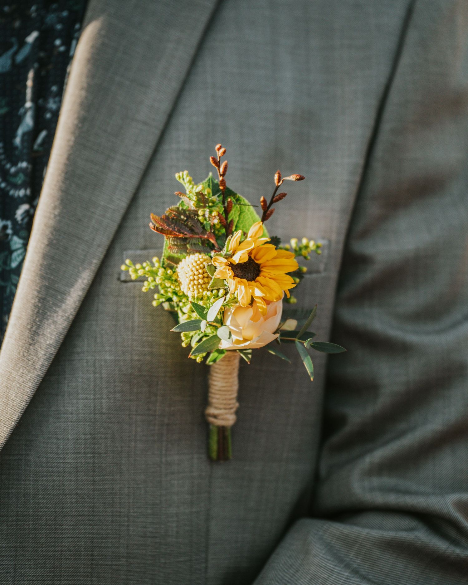 Three rustic boutonnieres with sunflowers, greenery and twine wrapped stems against a grey background.