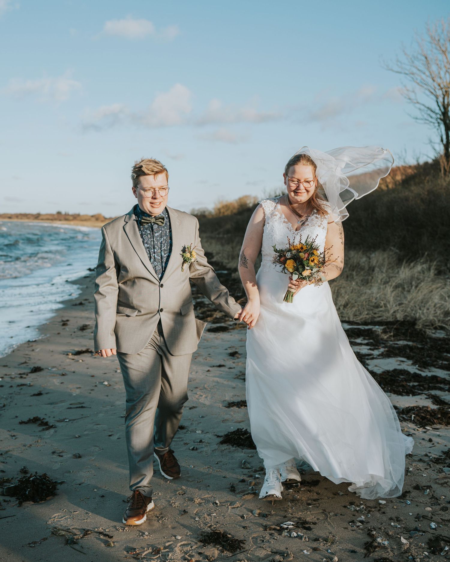 Couple in wedding attire walking hand-in-hand on a beach, bride in white gown with veil, groom in gray suit.
