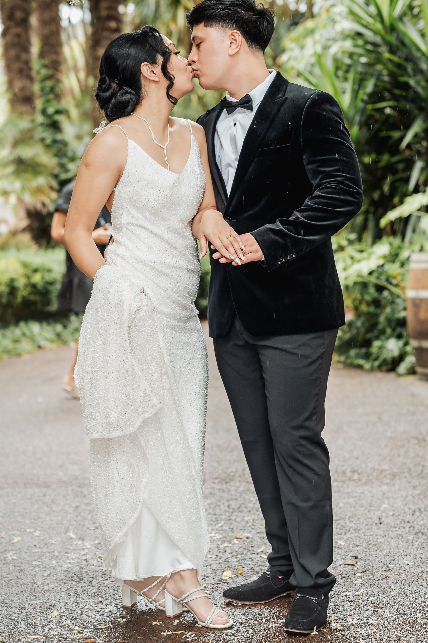 A romantic wedding couple share a kiss in a lush garden setting, bride in sparkly white gown and groom in velvet tuxedo.