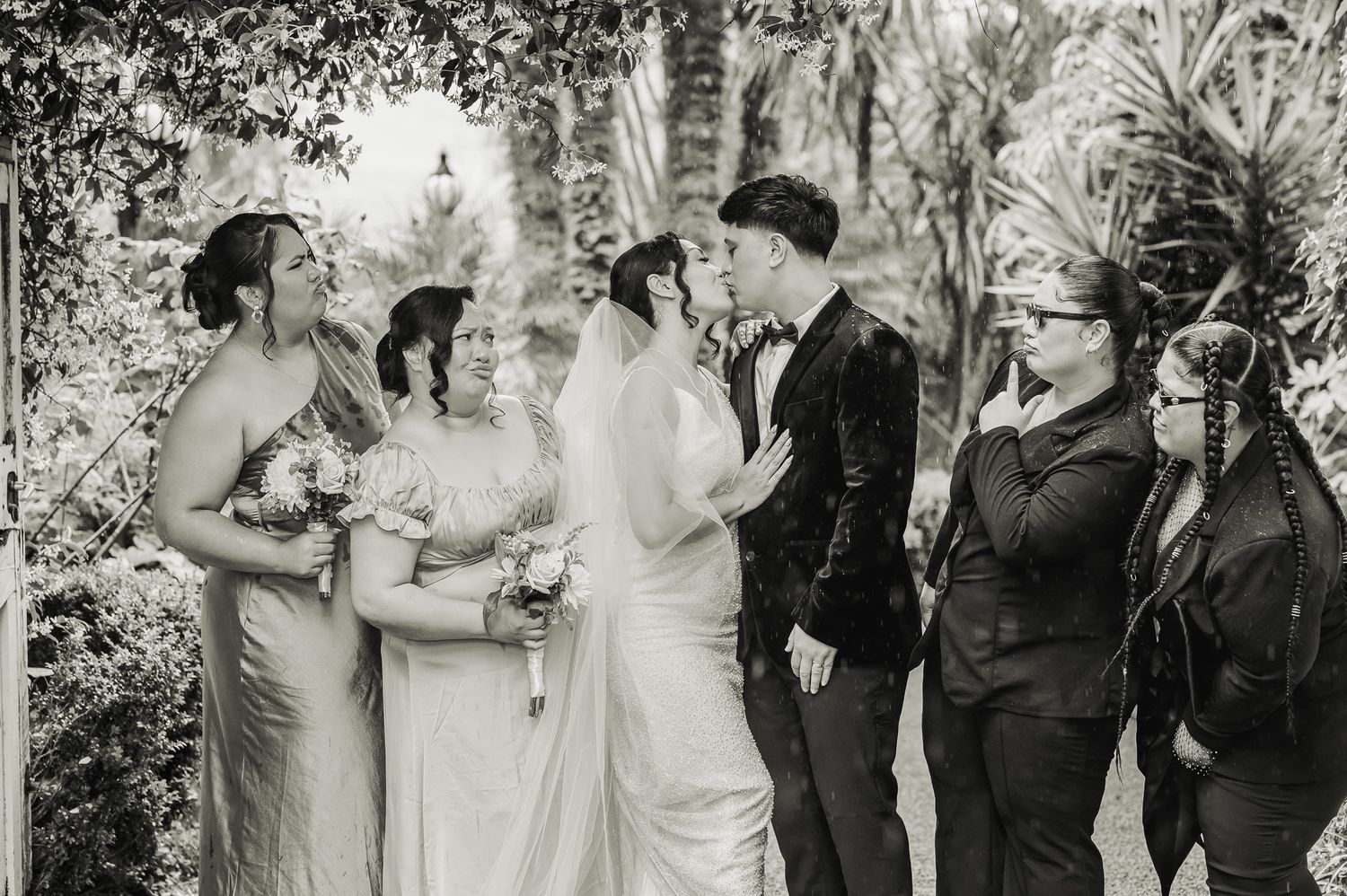 A vintage black and white photograph shows a group of wedding attendees gathered outdoors among lush foliage.