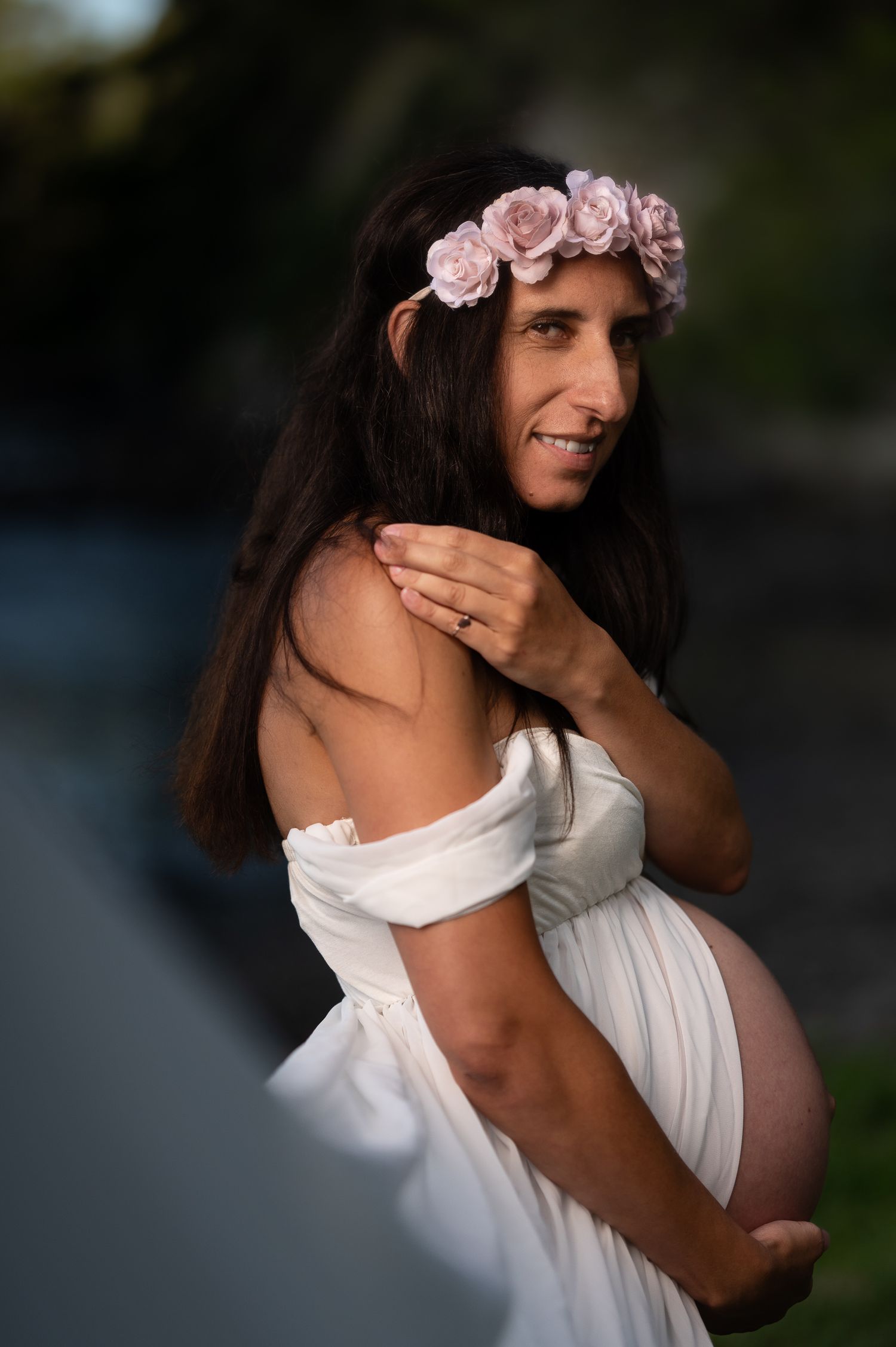 A pregnant person in a white off-shoulder dress and pink floral crown poses outdoors during golden hour lighting.
