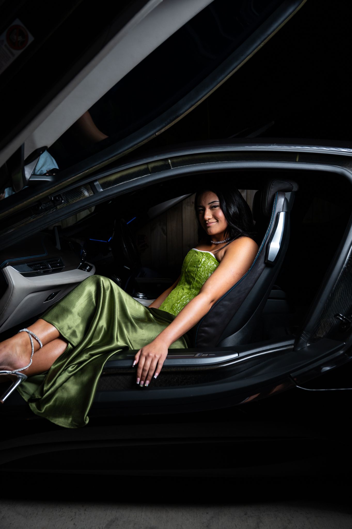 Young woman in elegant green satin dress posing inside luxury black car with door open.