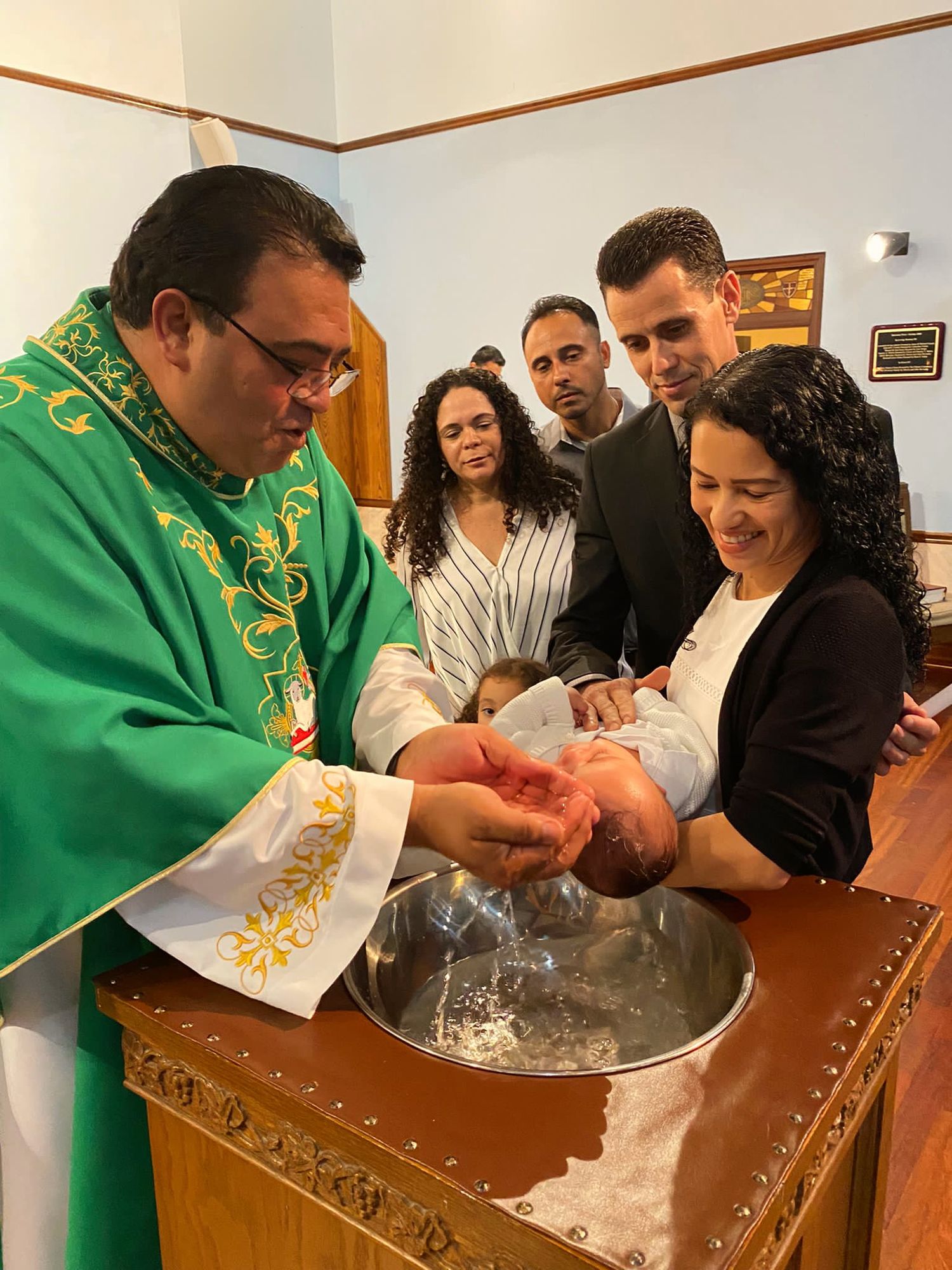 Religious ceremony with priest in green robes performing baptism ritual at wooden font.