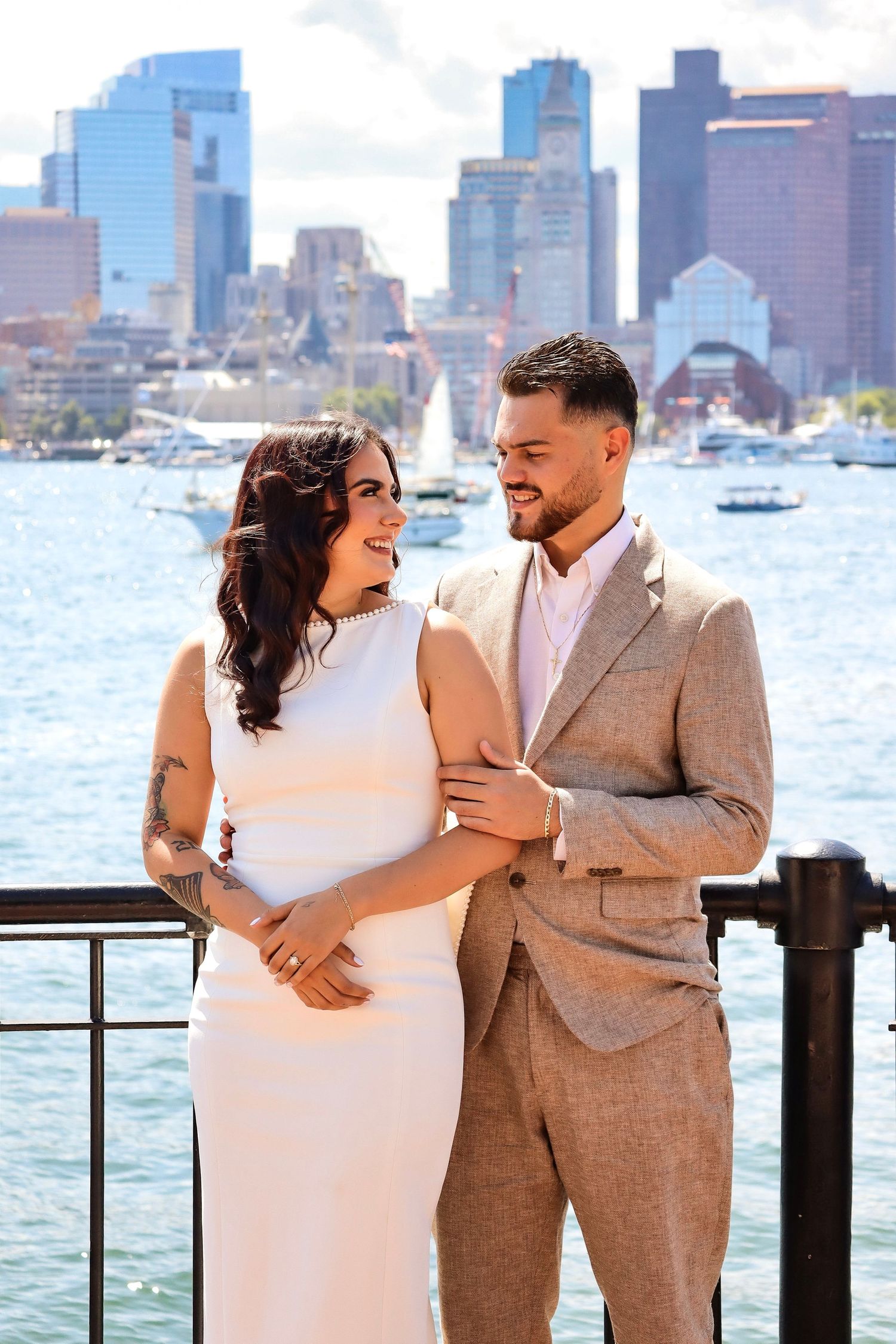 Couple poses along waterfront with Boston skyline visible in background.