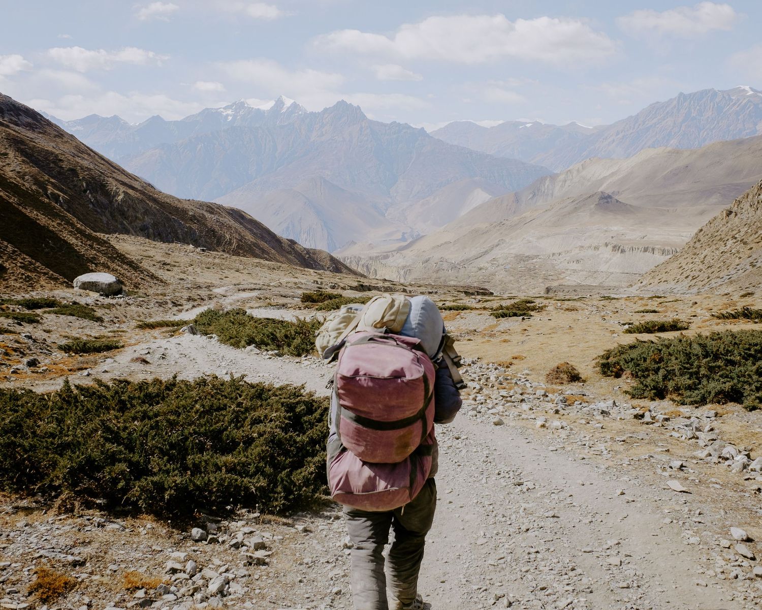 Trekkers carrying gear traverse a high-altitude mountain path with panoramic views.