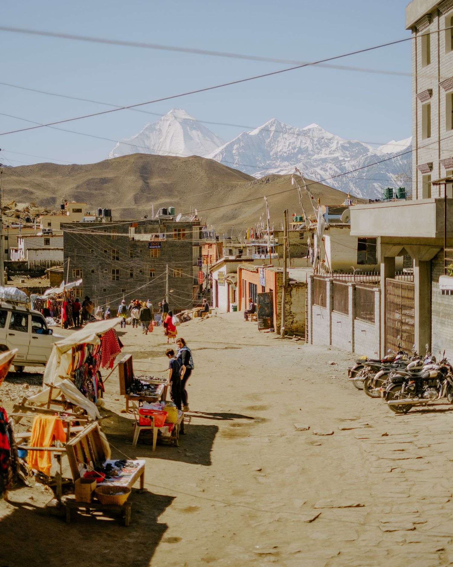 A panoramic view of a mountain village marketplace with vendors displaying goods along dusty streets against snowy peaks.