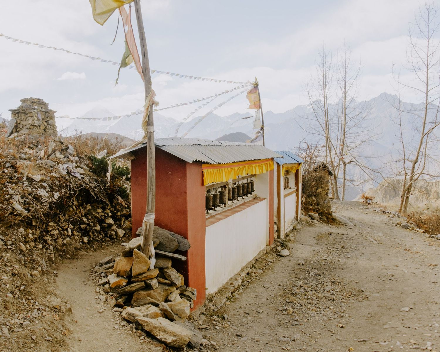 Stone walls and structures line a mountain path with prayer flags and distant peaks in the background.