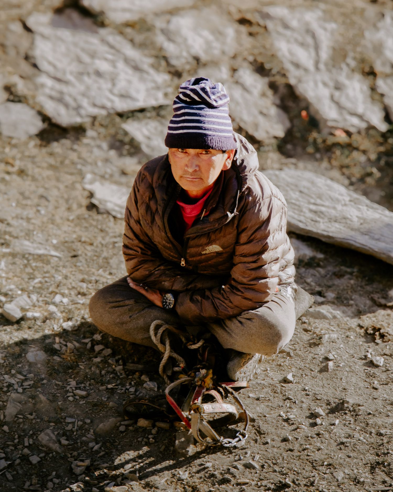 A person in a brown jacket and striped cap crouches on rocky terrain while exploring outdoors.