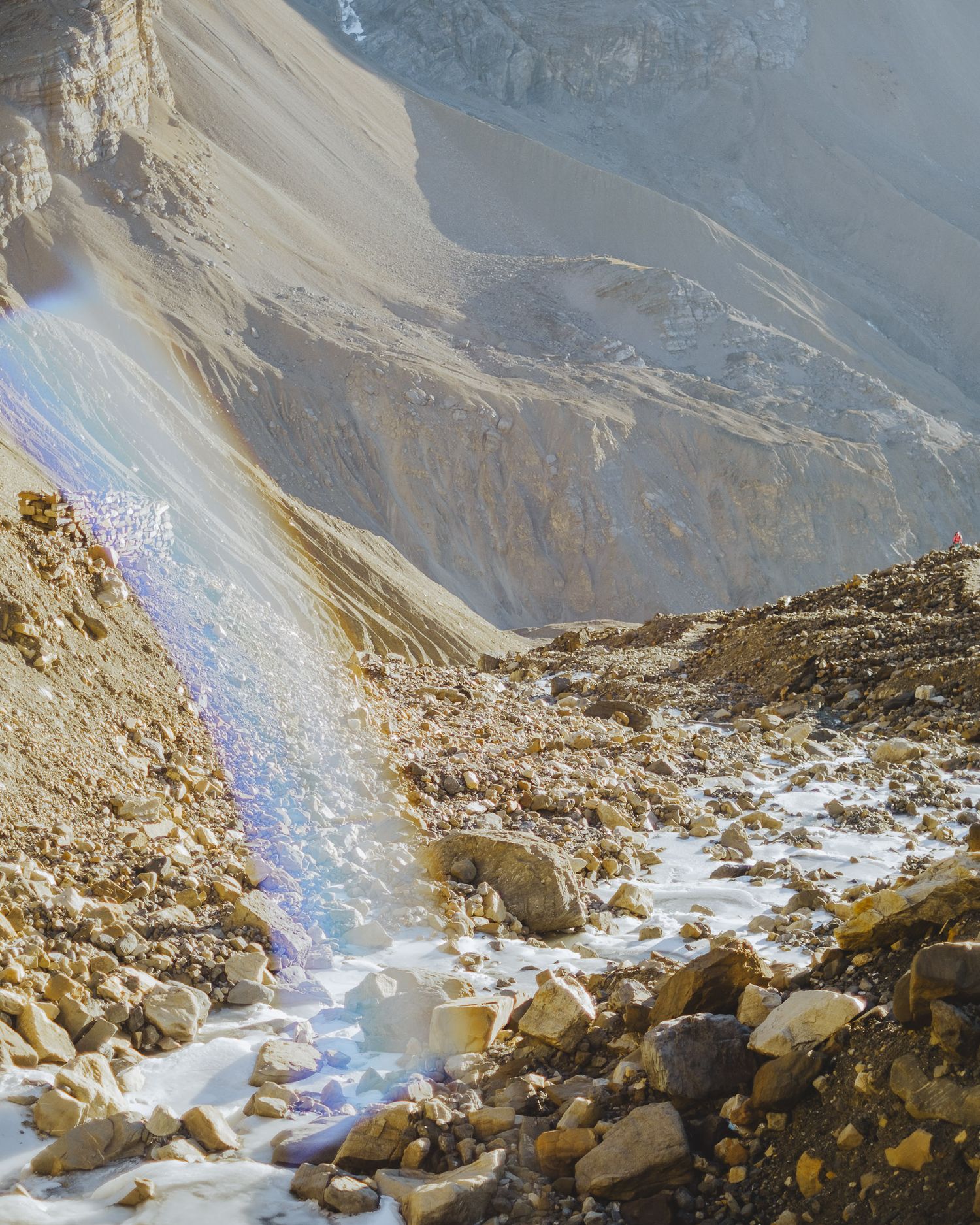 Rocky mountain trail with scattered snow and a rainbow lens flare against steep cliffs.